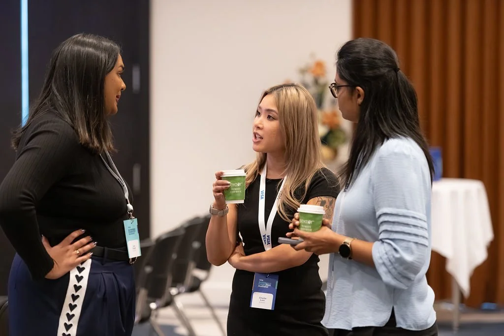 Three women engaged in a conversation at a conference, holding coffee cups.