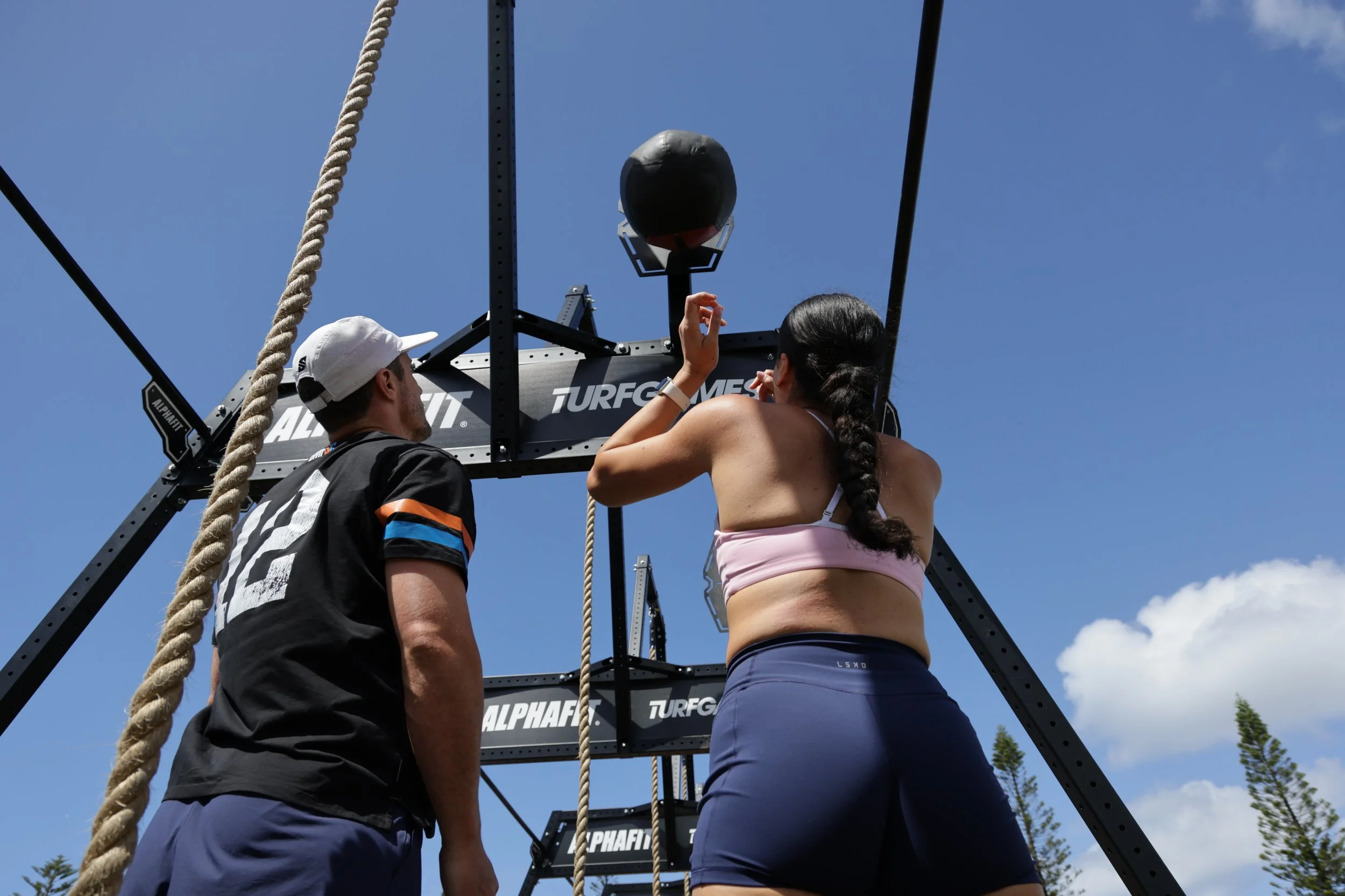 A woman in athletic clothing is preparing to participate in a fitness competition with a man assisting her, standing near a rig that has a medicine ball and climbing ropes, outdoors on a sunny day.