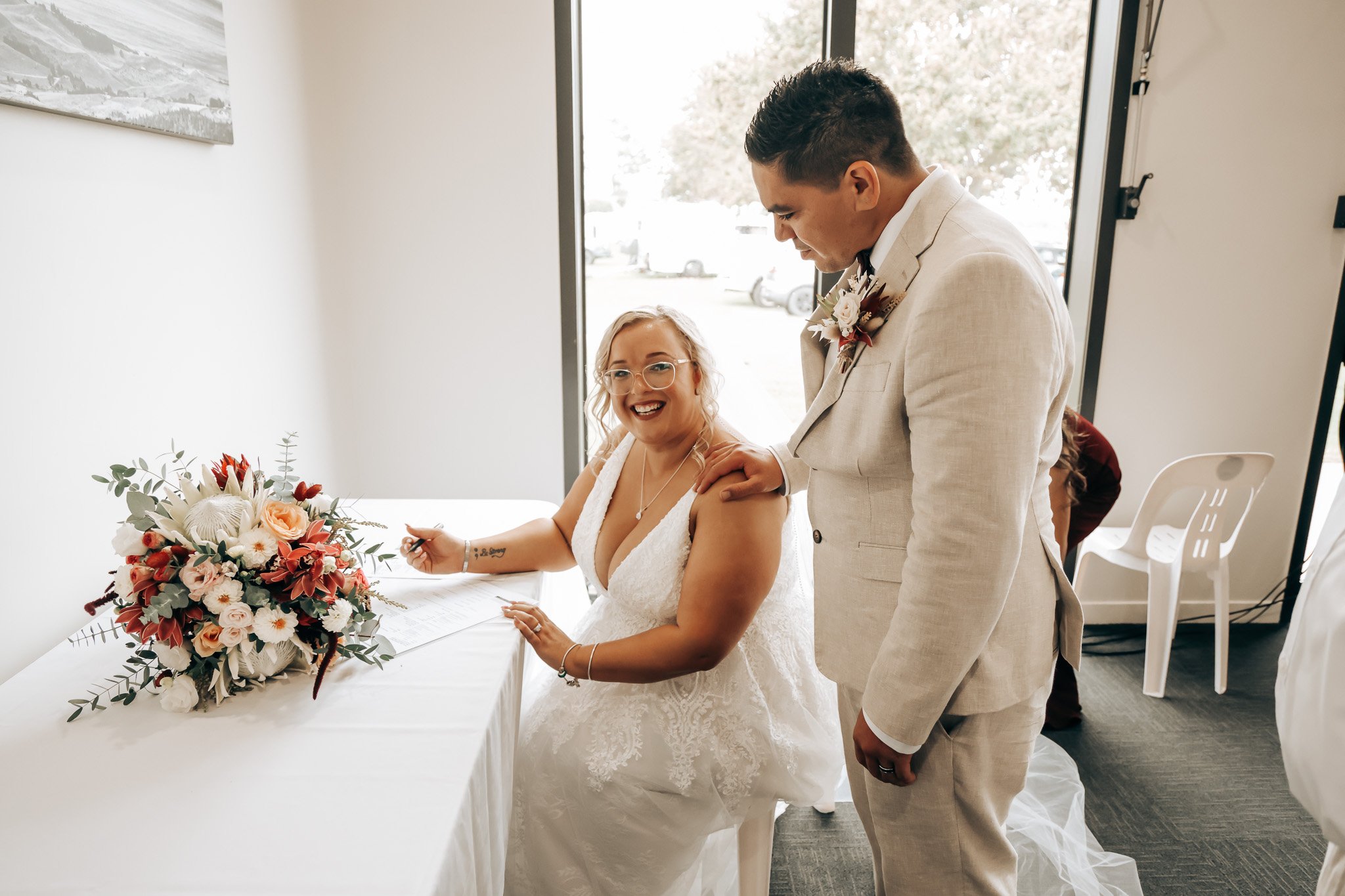 A bride and groom signing marriage documents during their wedding ceremony, with a floral bouquet on the table.