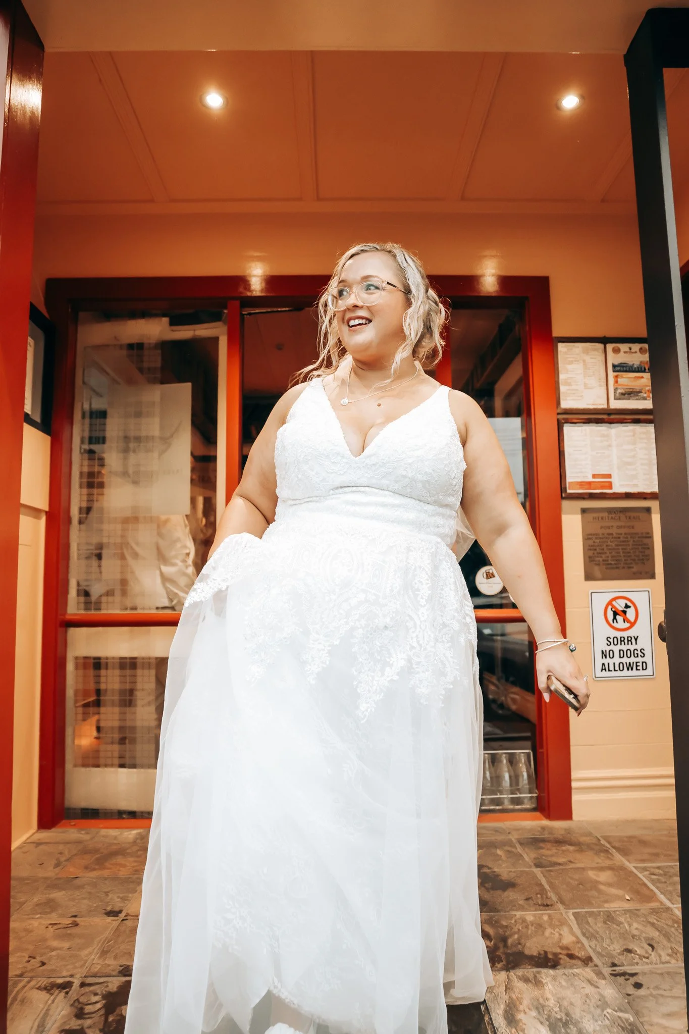 A woman in a white wedding dress standing inside a building, smiling and holding a phone in her right hand, with a restaurant or shop entrance behind her.