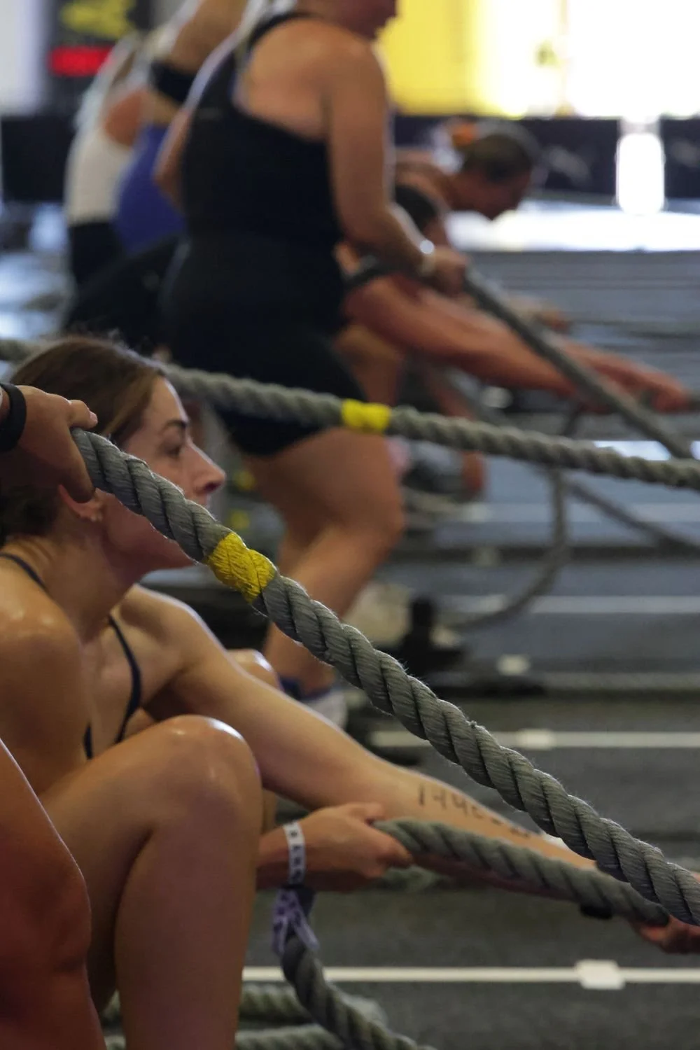 Women with athletic gear participating in a fitness class using battle ropes at a gym, some stretching and others sitting on the floor, focused on the workout.