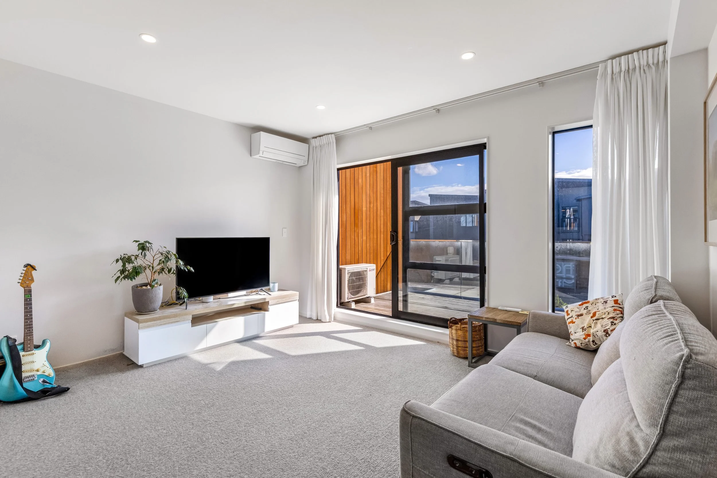 A modern living room with a gray sofa, flat-screen TV on a white stand, a blue electric guitar, a potted plant, and floor-to-ceiling windows with white curtains leading to a balcony with a wood-paneled wall and an outdoor air conditioning unit.