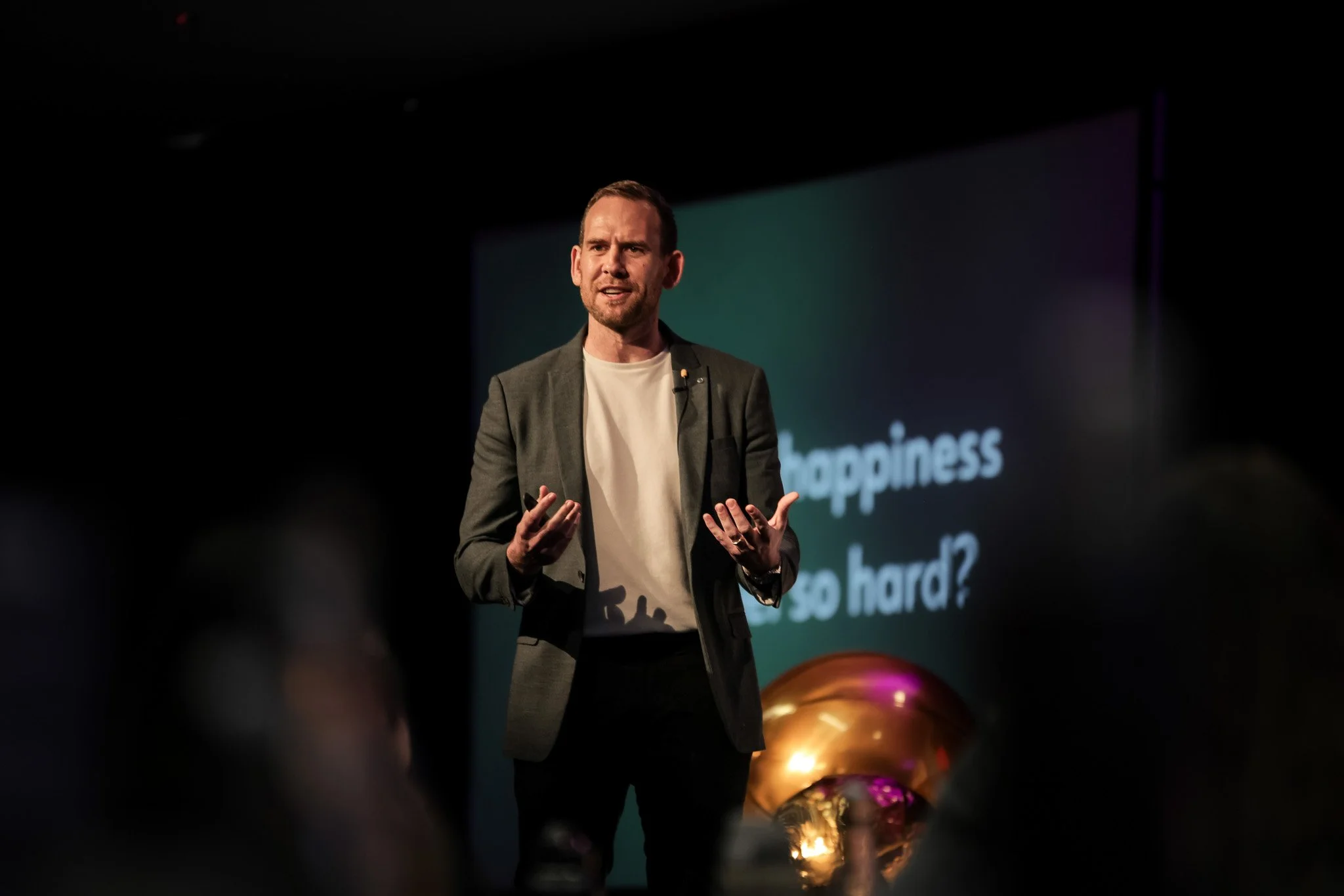 A man in a blazer and t-shirt speaking during a presentation on stage, with text on a screen behind him that says 'happiness' and part of another word.