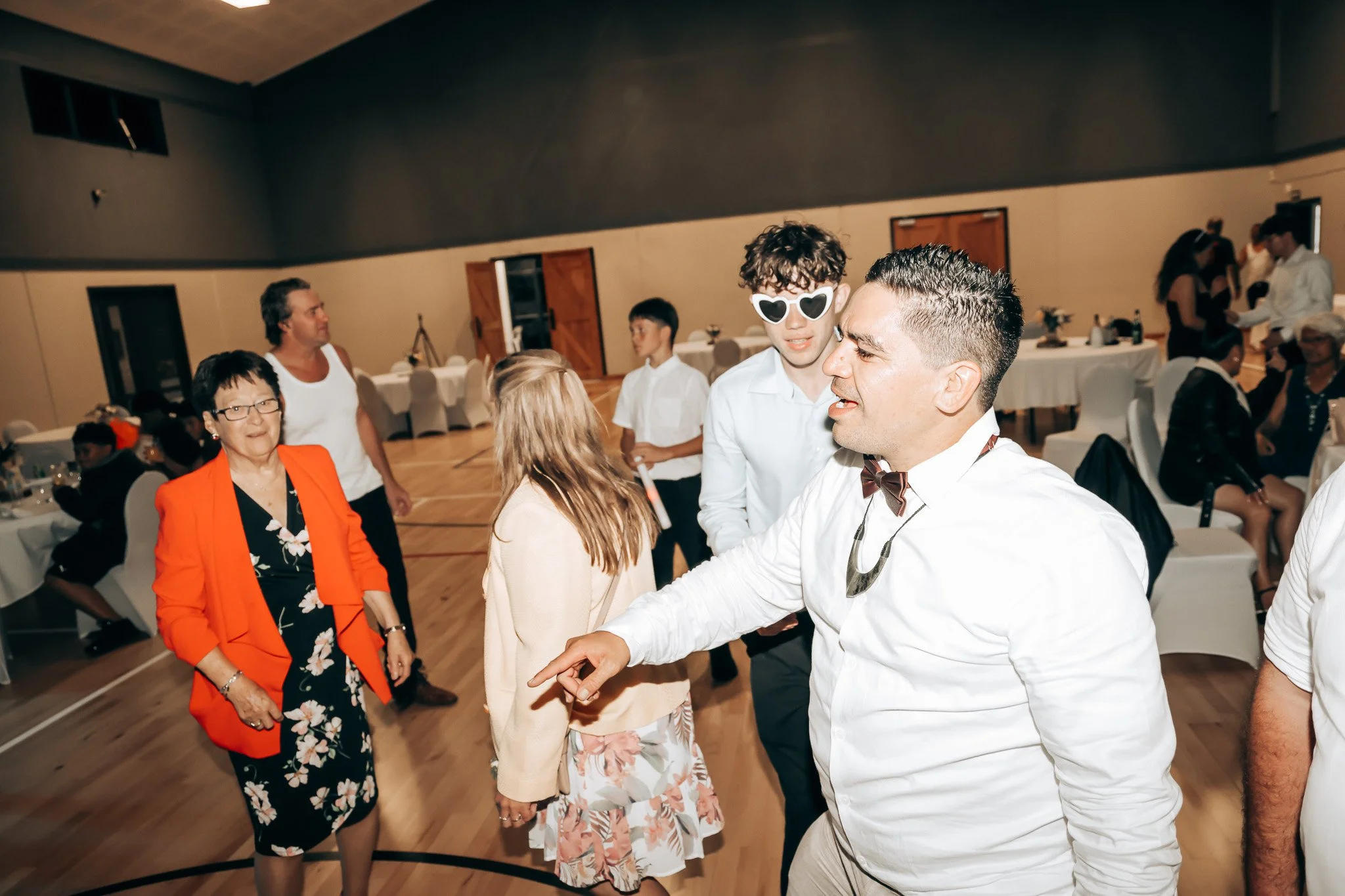 People dancing and socializing at a party or celebration in a banquet hall with decorated tables and chairs.