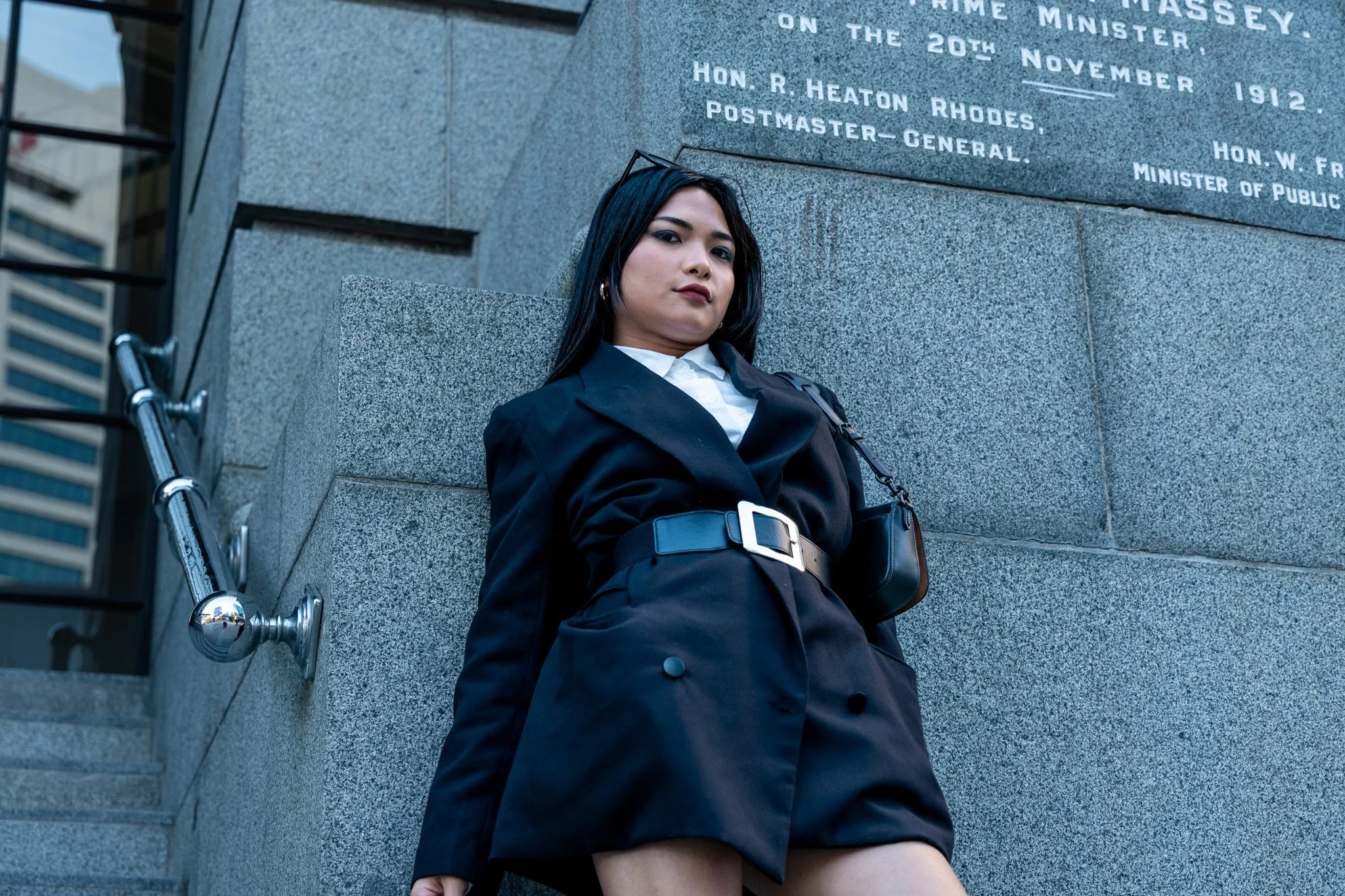 A woman with dark hair, wearing a black blazer and belt, leaning against a gray stone wall with engraved text. A walker is partially visible in the foreground.