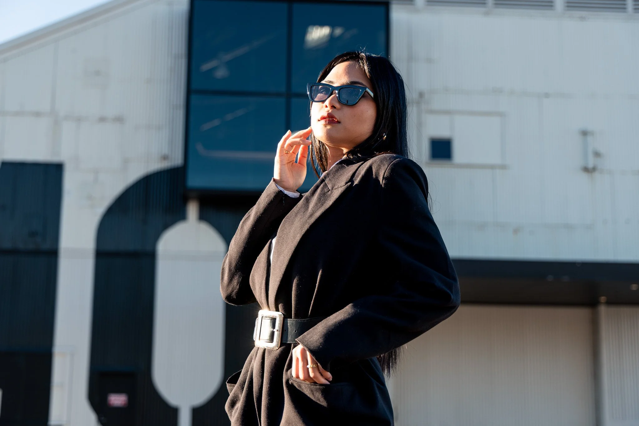 A woman with long dark hair and sunglasses wearing a black blazer, standing outdoors near a modern building with large windows and a curved roof.