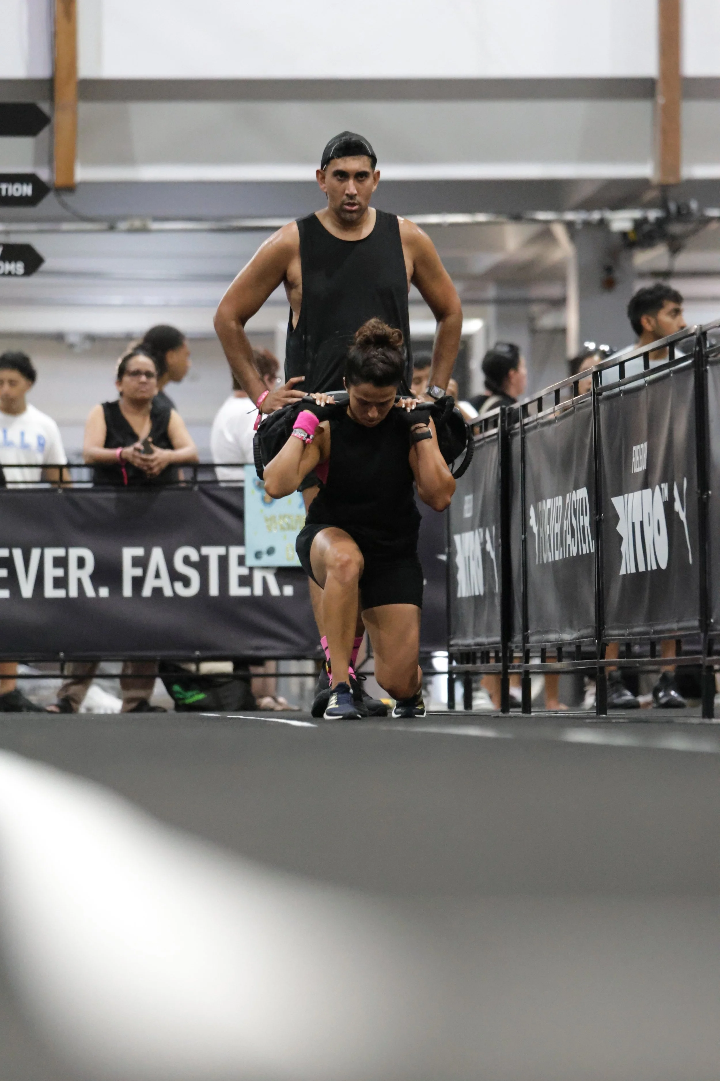 A man and woman in athletic clothing working out at an indoor gym, with the woman doing lunges while the man stands behind her. There are people and gym equipment in the background.