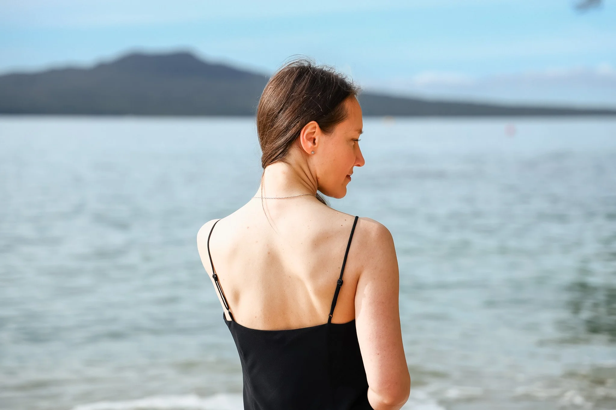 A woman with brown hair and fair skin standing on a beach with water and mountains in the background, wearing a black dress with thin straps.