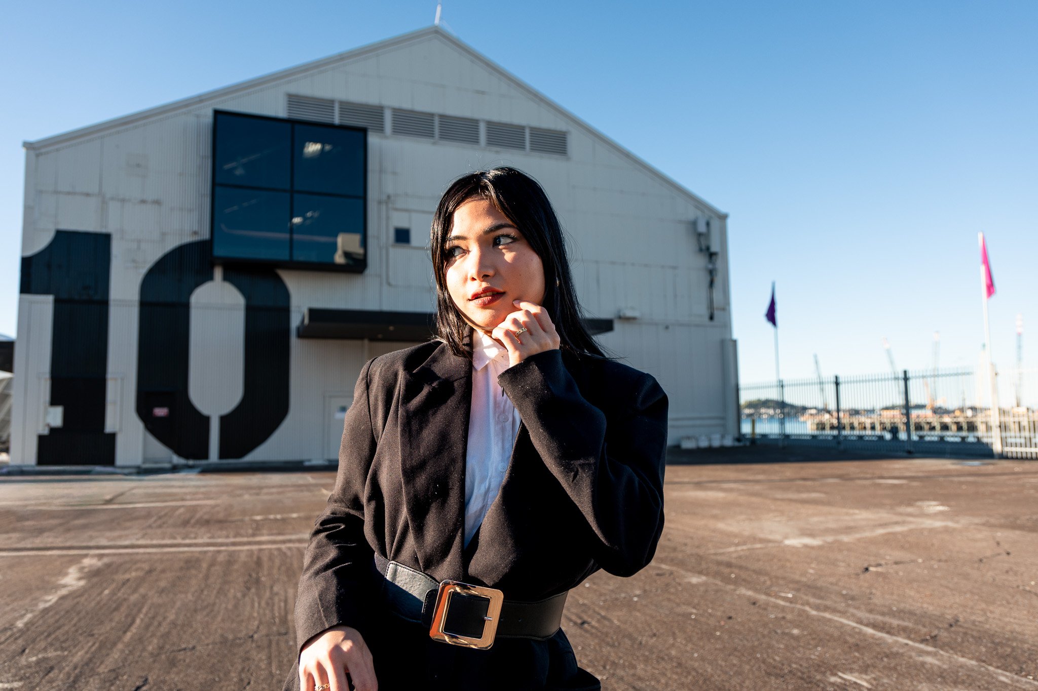 A young woman with black hair and a thoughtful expression, standing outdoors in front of a large white building with a black letter 'O' on it, wearing a black blazer, white shirt, and a wide belt with a large square buckle.