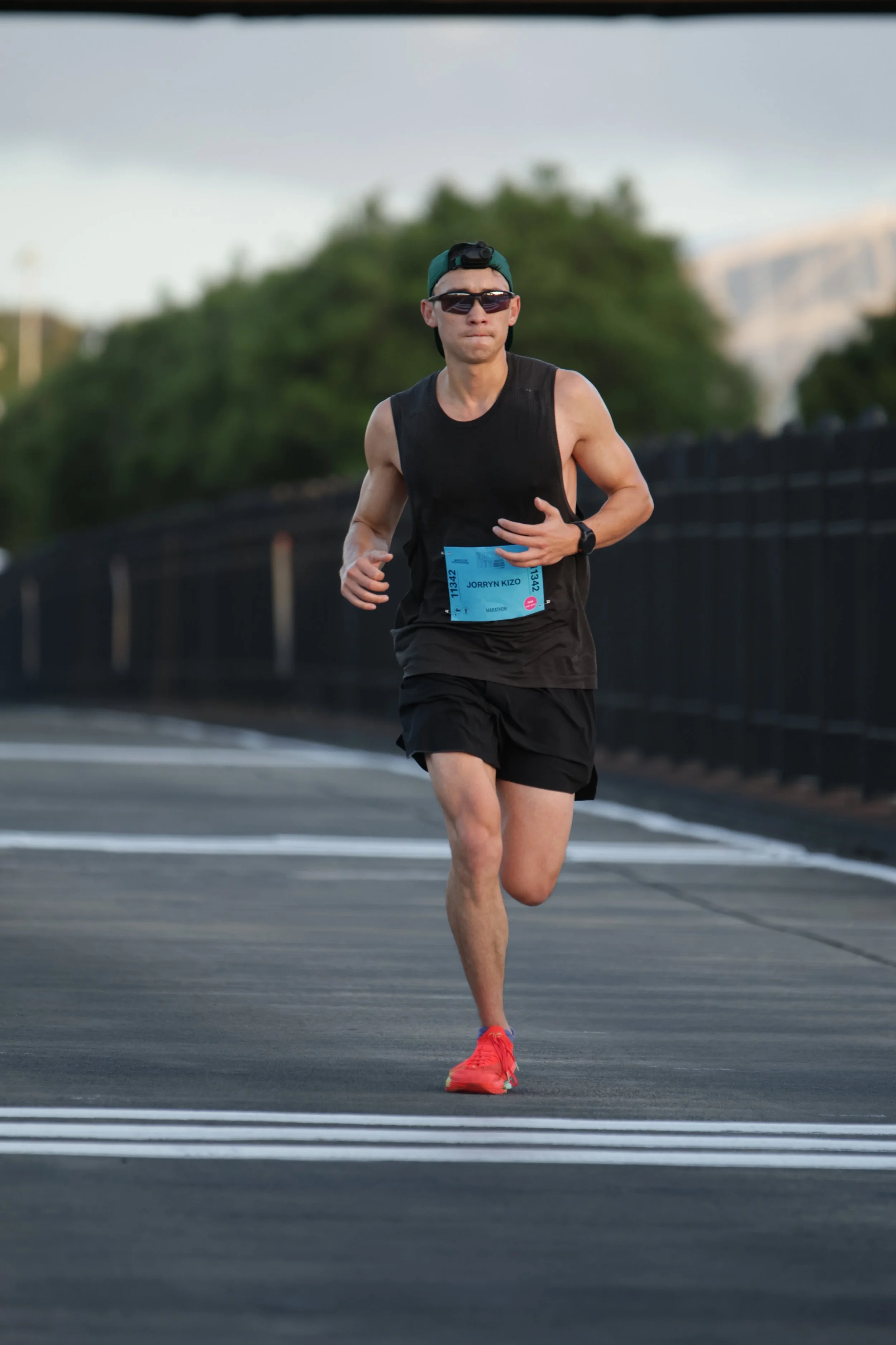 Male runner in black athletic wear, wearing sunglasses, a green cap worn backwards, and bright orange running shoes, participating in a race on a paved outdoor road.