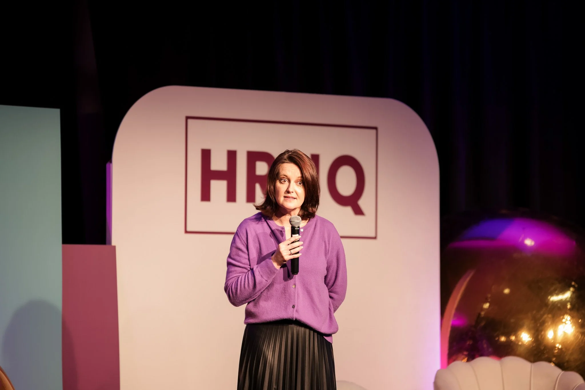 A woman speaking into a microphone on stage during an event with a large HBO Q logo in the background.