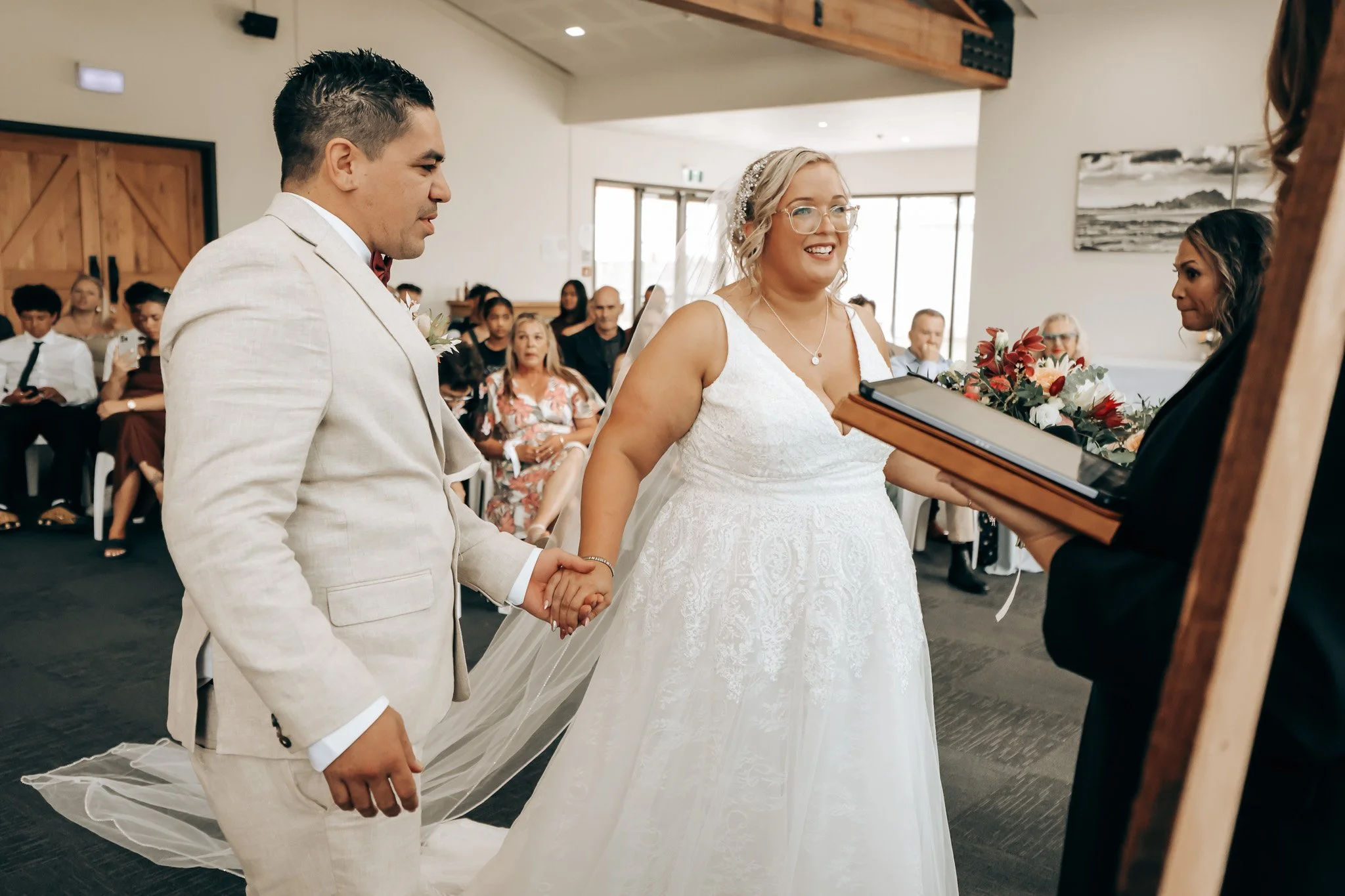 A couple is holding hands during a wedding ceremony, with the bride smiling and the groom looking serious, surrounded by seated guests in a bright, modern venue.