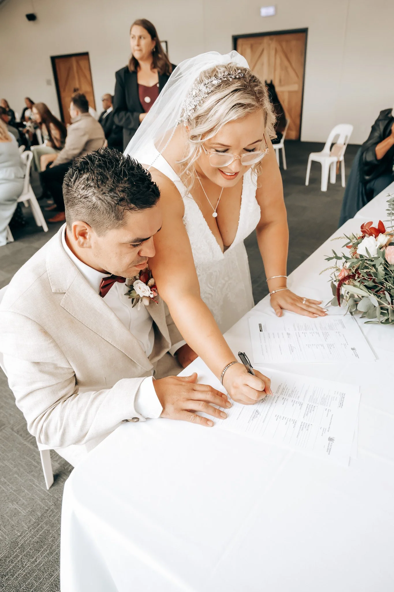 A bride and groom signing a marriage certificate at a wedding ceremony with guests in the background.