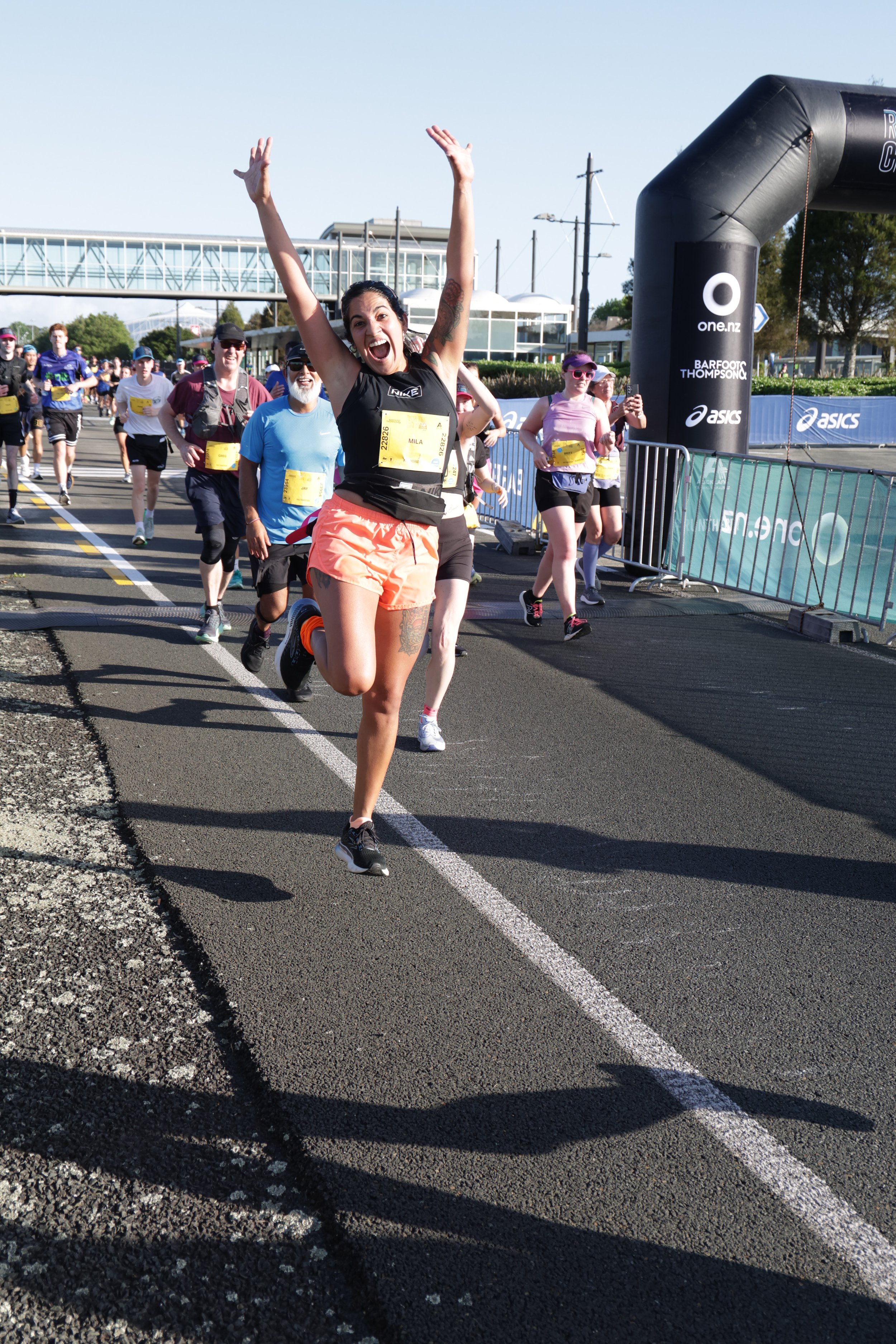 A group of runners crossing the finish line at a race, with a woman in front celebrating with her arms raised and a wide smile.