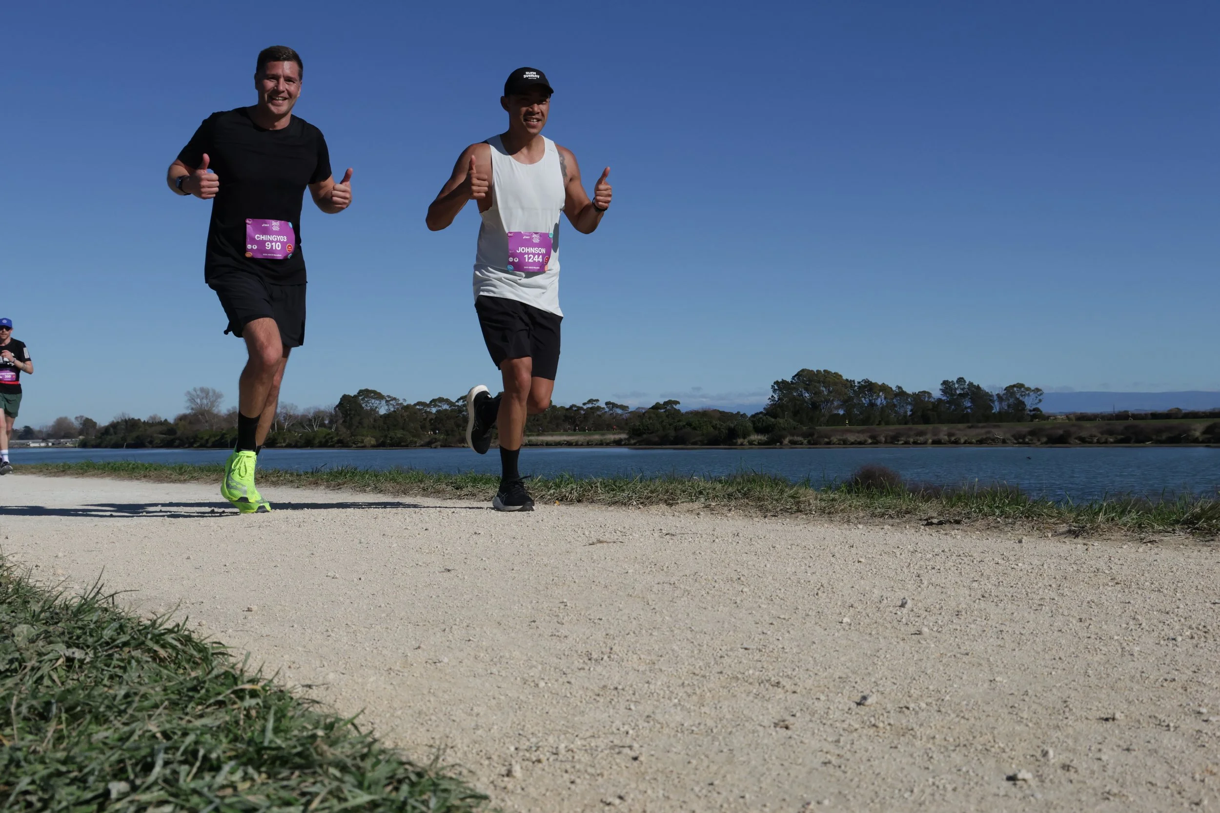 Two men running outdoors near a body of water with a clear blue sky, one in a black shirt and the other in a white tank top, both giving thumbs up and wearing race bibs.
