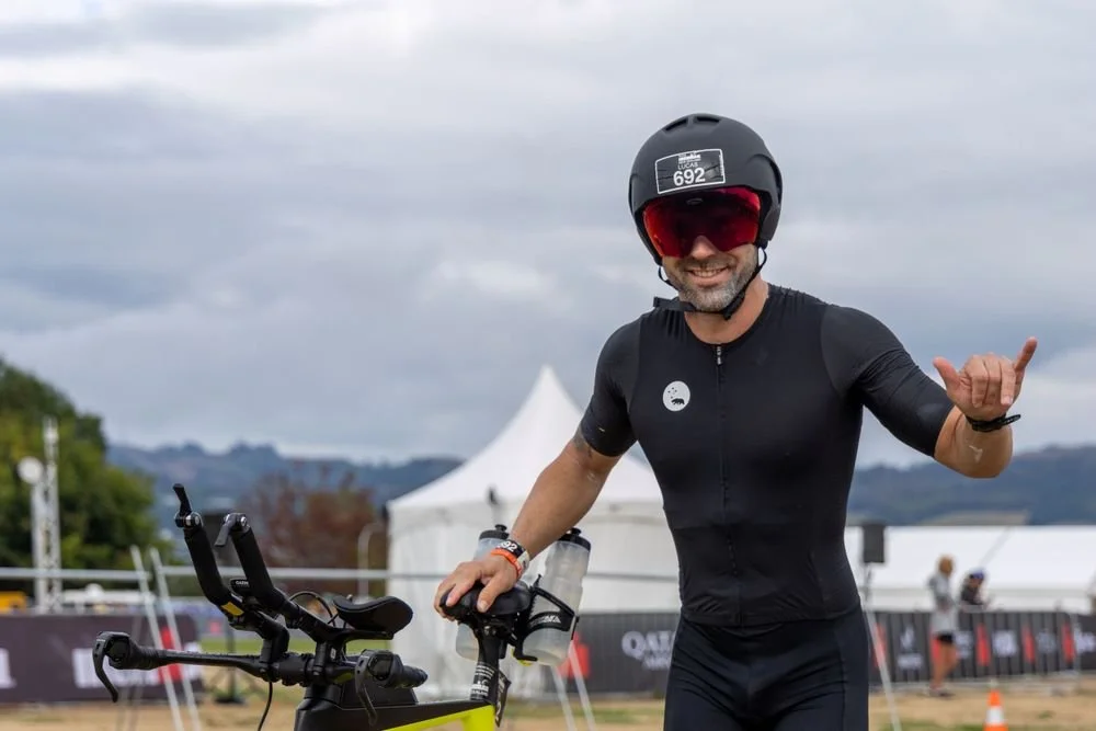 A smiling man wearing a black cycling helmet, sunglasses, and black athletic outfit, making a shaka hand gesture while standing beside his bike at an outdoor event.
