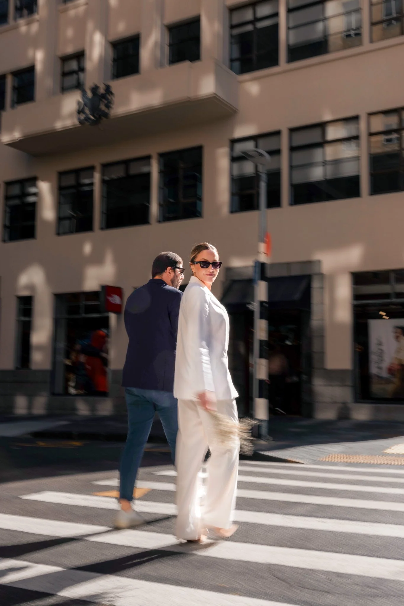 A woman dressed in a white suit and sunglasses crossing a city street at a crosswalk, with a man in a dark blazer beside her, behind her is a beige building with multiple windows and a streetlamp.