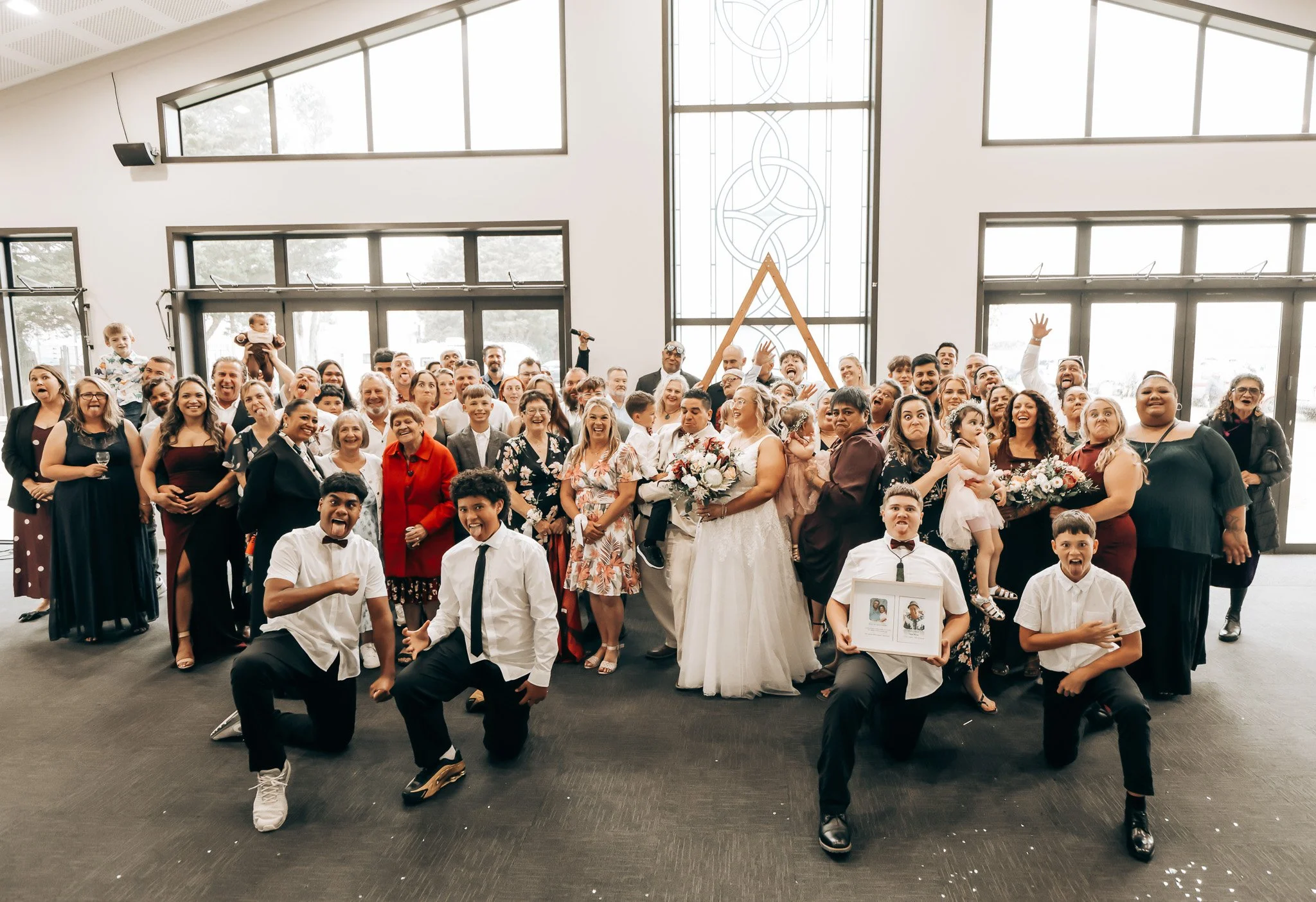 Group of wedding guests and children posing for a photo inside a modern venue with large windows and decorative glass wall, some smiling and making funny faces.
