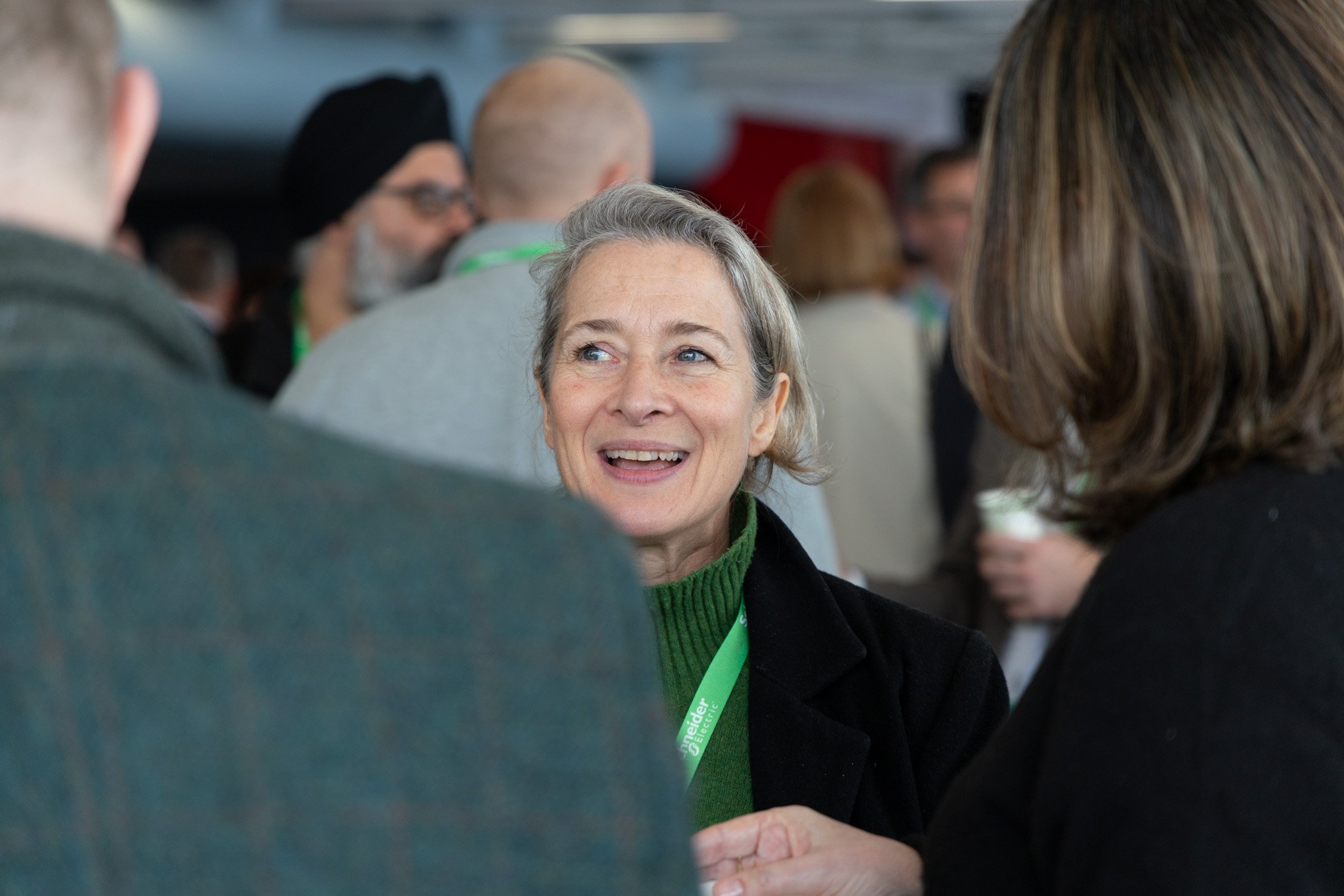 A woman with short gray hair and a green sweater smiling and talking to another woman with long brown hair. There are people in the background at a busy event or conference.