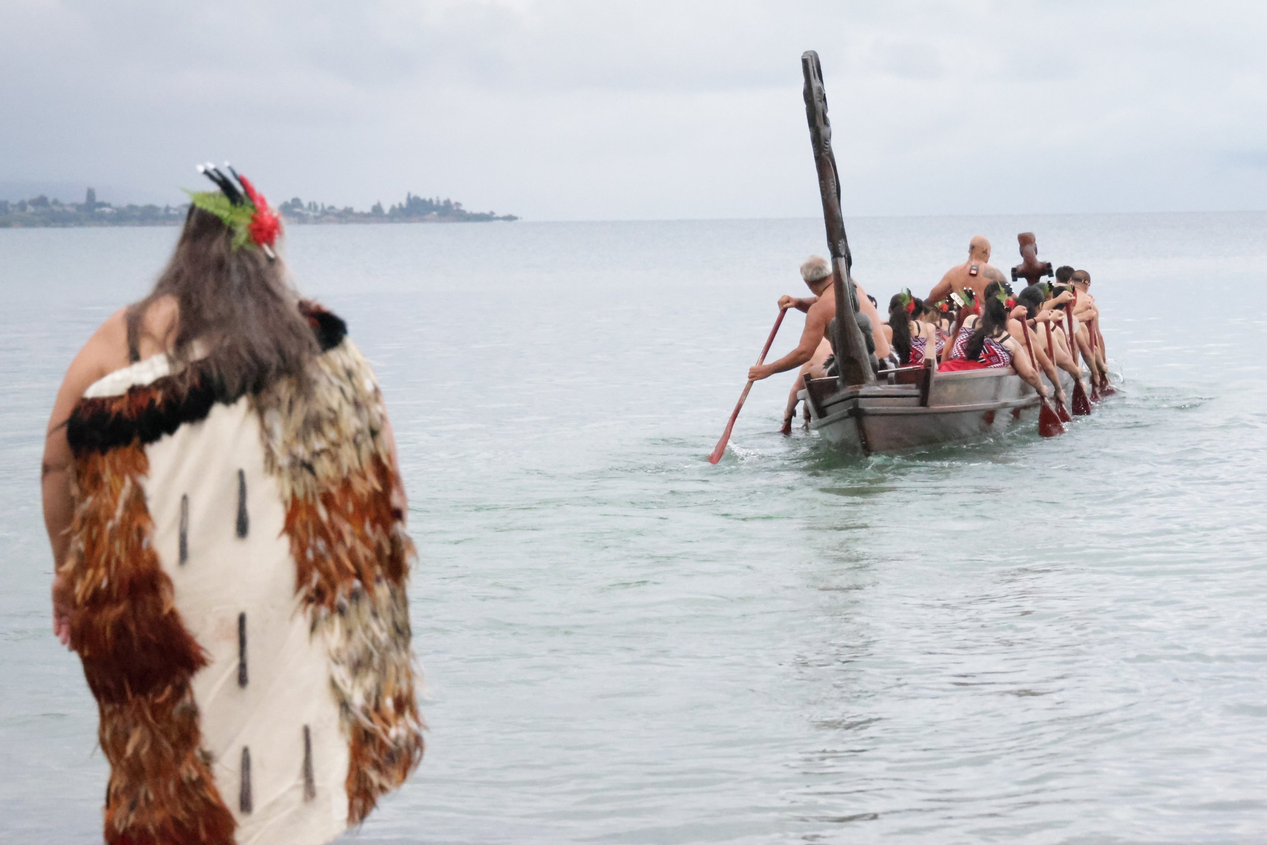 A person in traditional Native American attire with feathered and fur adornments observing a canoe filled with people paddling on a calm body of water under an overcast sky.