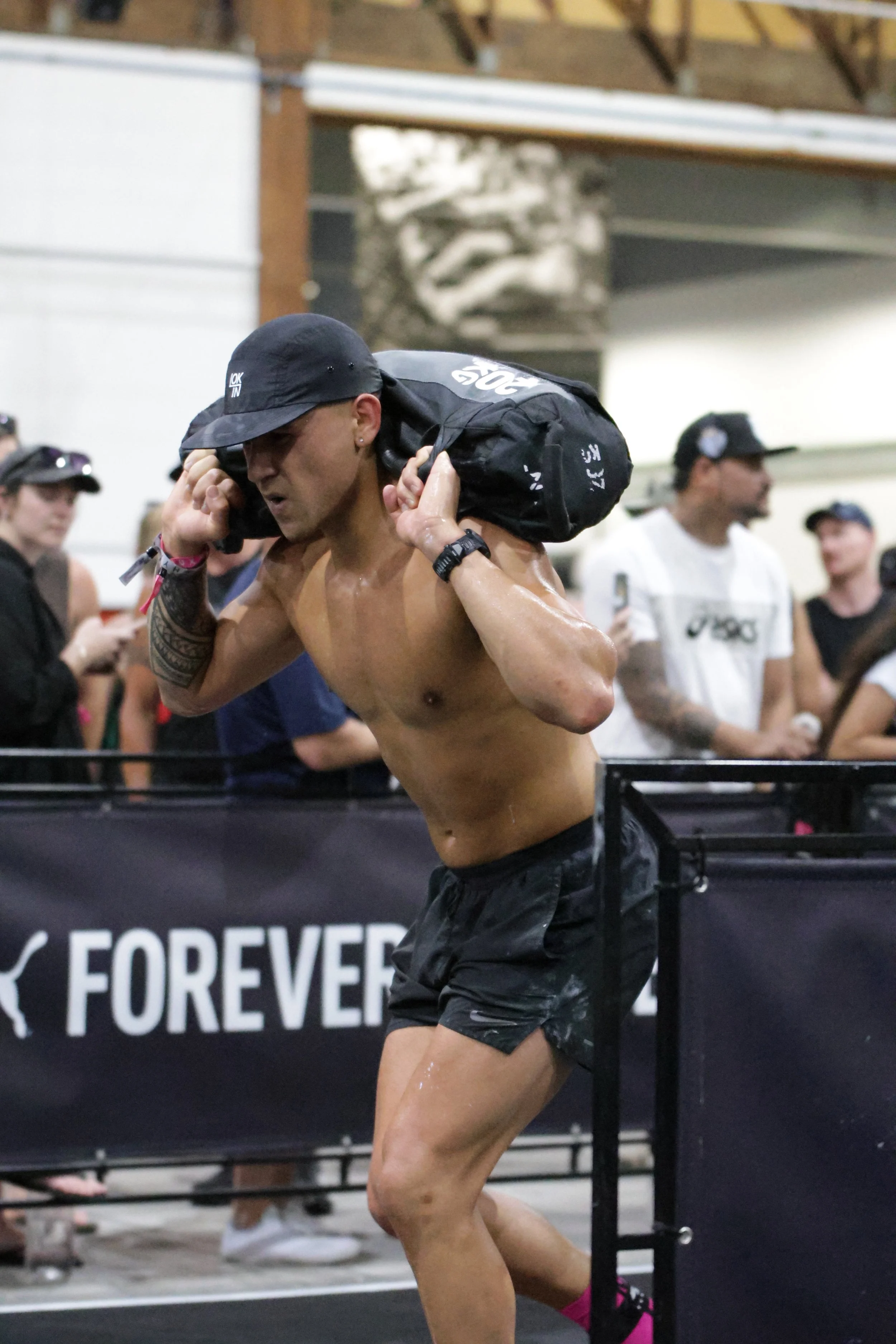 A shirtless man with a black cap carrying a black bag over his shoulder at a fitness event, with several people watching in the background.