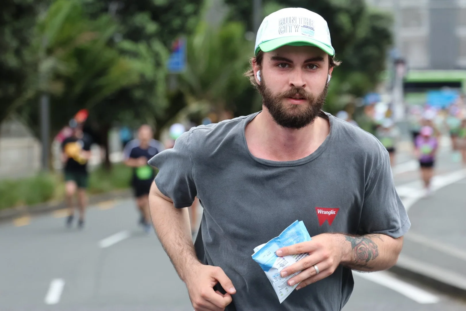 A man with a beard and tattoos running in a race, holding a blue packet, wearing a gray t-shirt and a white cap with green trim, with other runners and trees in the background.