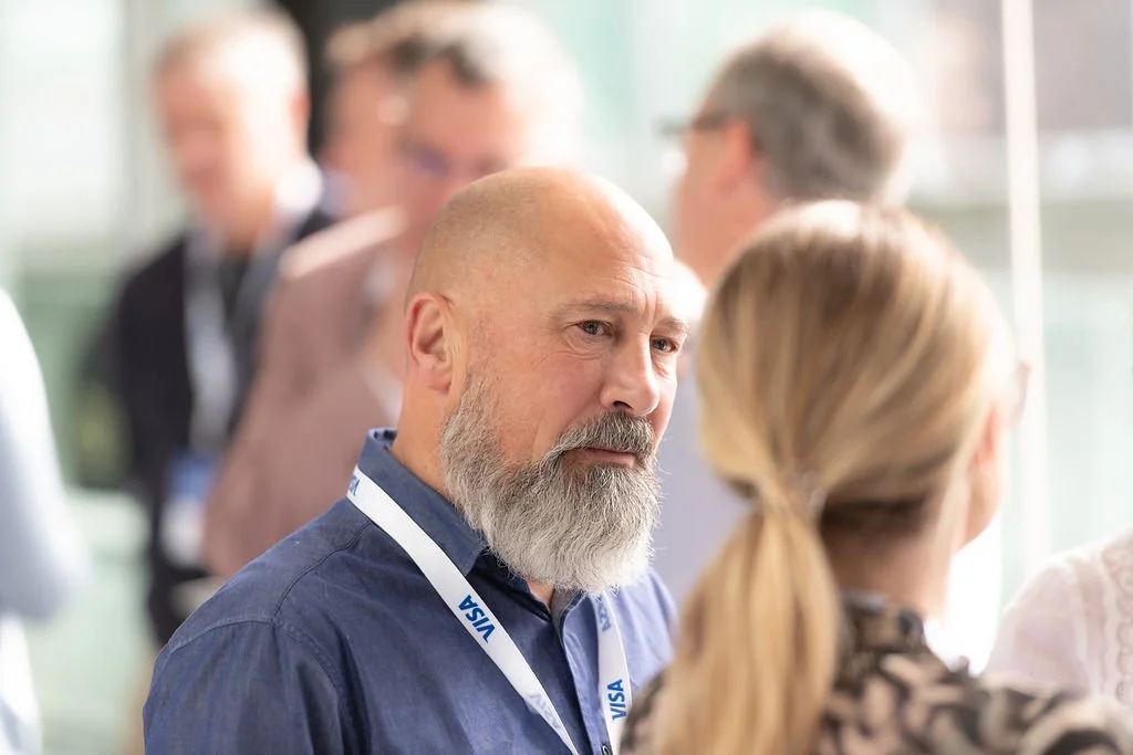 A man with a gray beard and blue shirt talking to a woman with blonde hair at a conference, with other attendees blurred in the background.
