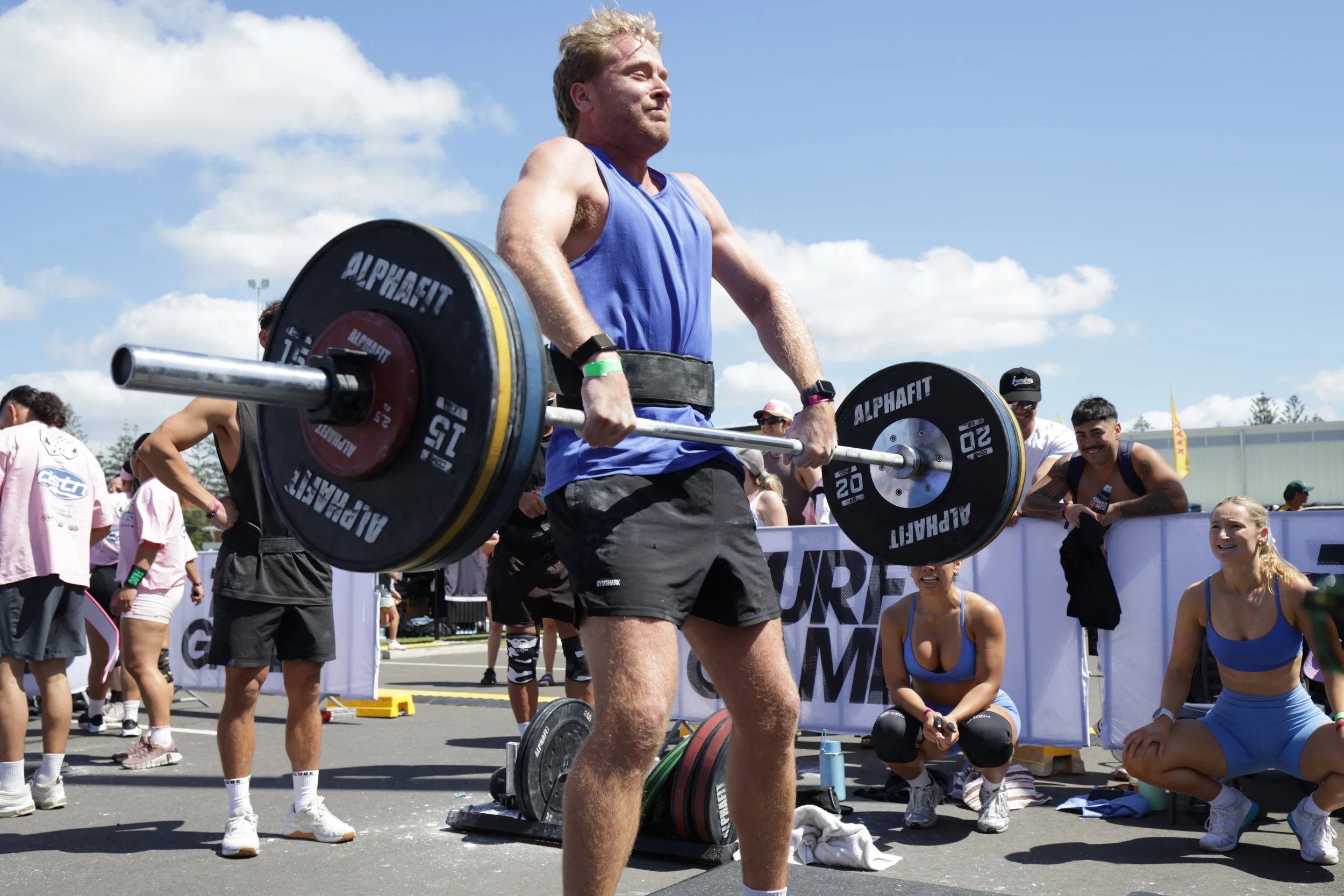 A man in a blue tank top and black shorts lifting a barbell with weights during a CrossFit competition outdoors. Several spectators and participants are watching, with some sitting and others standing. The sky is clear with some clouds.