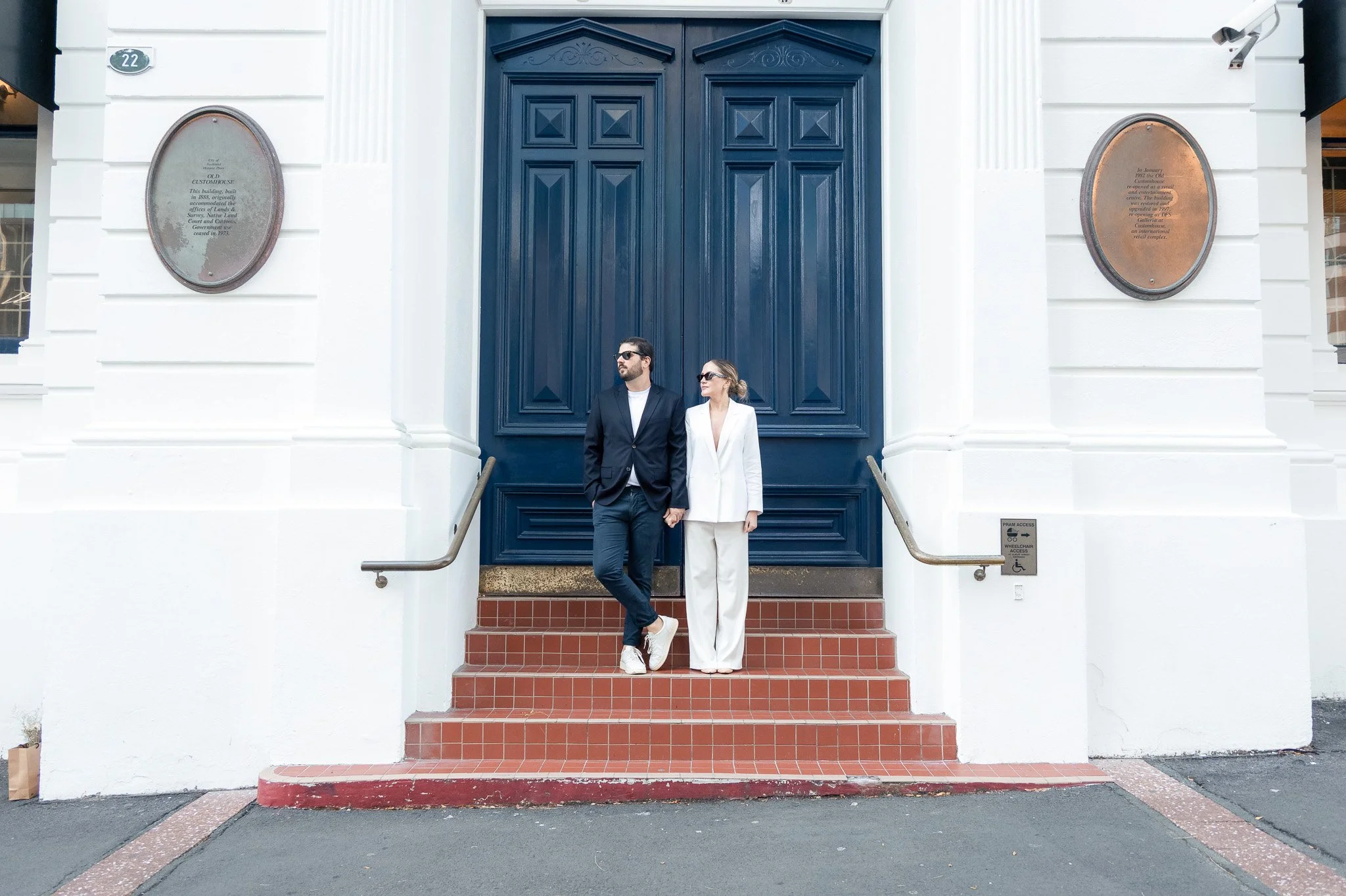 A man and a woman standing in front of a large blue door on white building exterior, holding hands and looking to the side, dressed in formal attire.