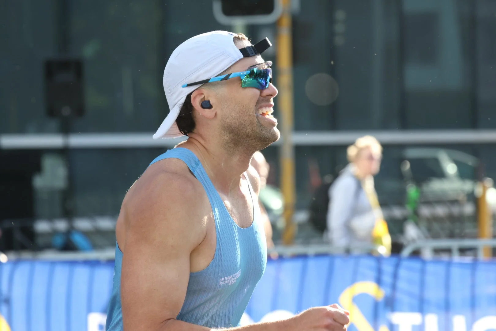 A male marathon runner wearing a white cap backward, sunglasses, and a blue sleeveless shirt, smiling as he runs in an outdoor race on a sunny day.