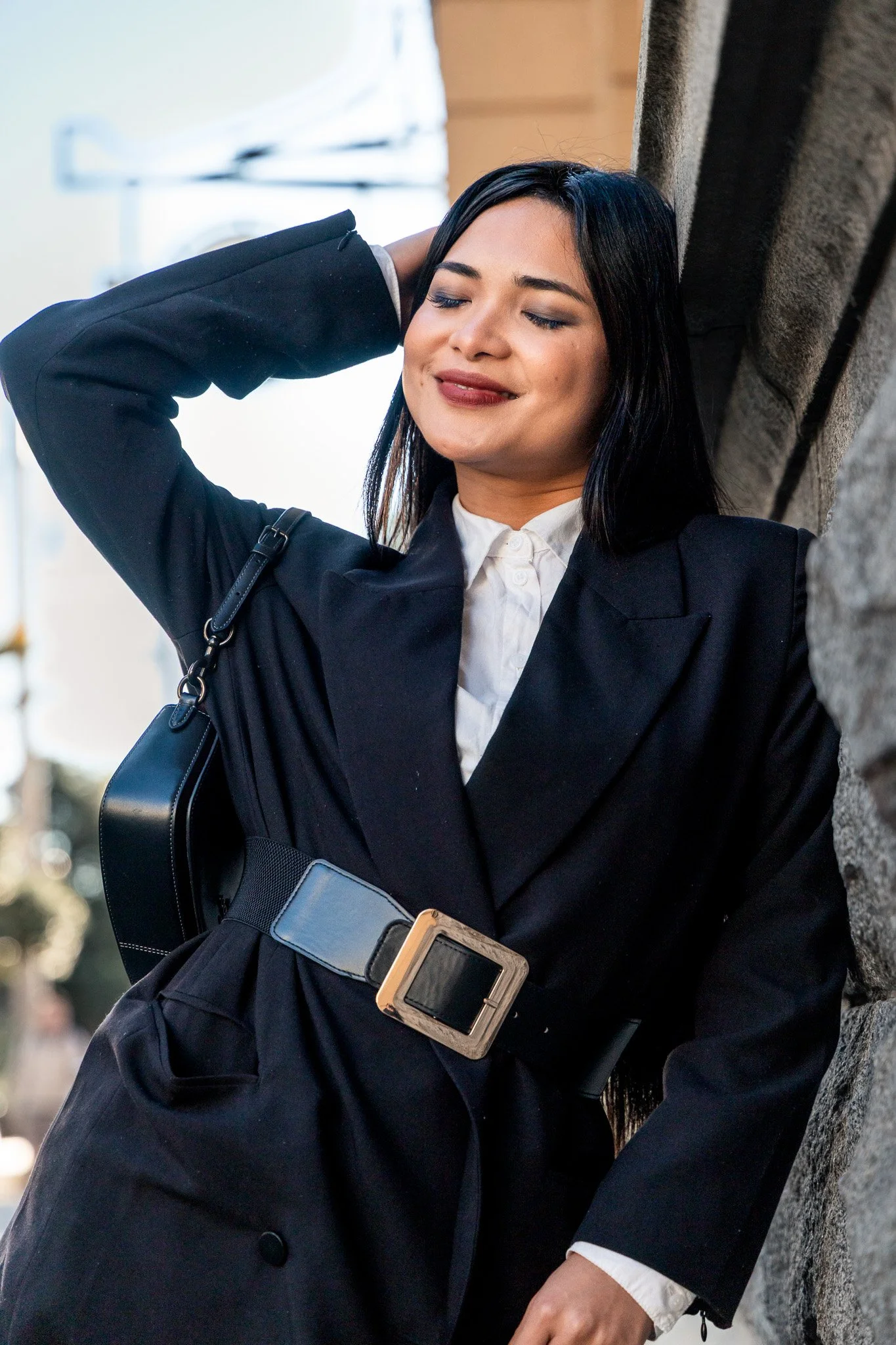 A woman with black hair, wearing a black blazer and a white shirt, leaning against a stone wall with her eyes closed, smiling softly, and one hand resting behind her head.