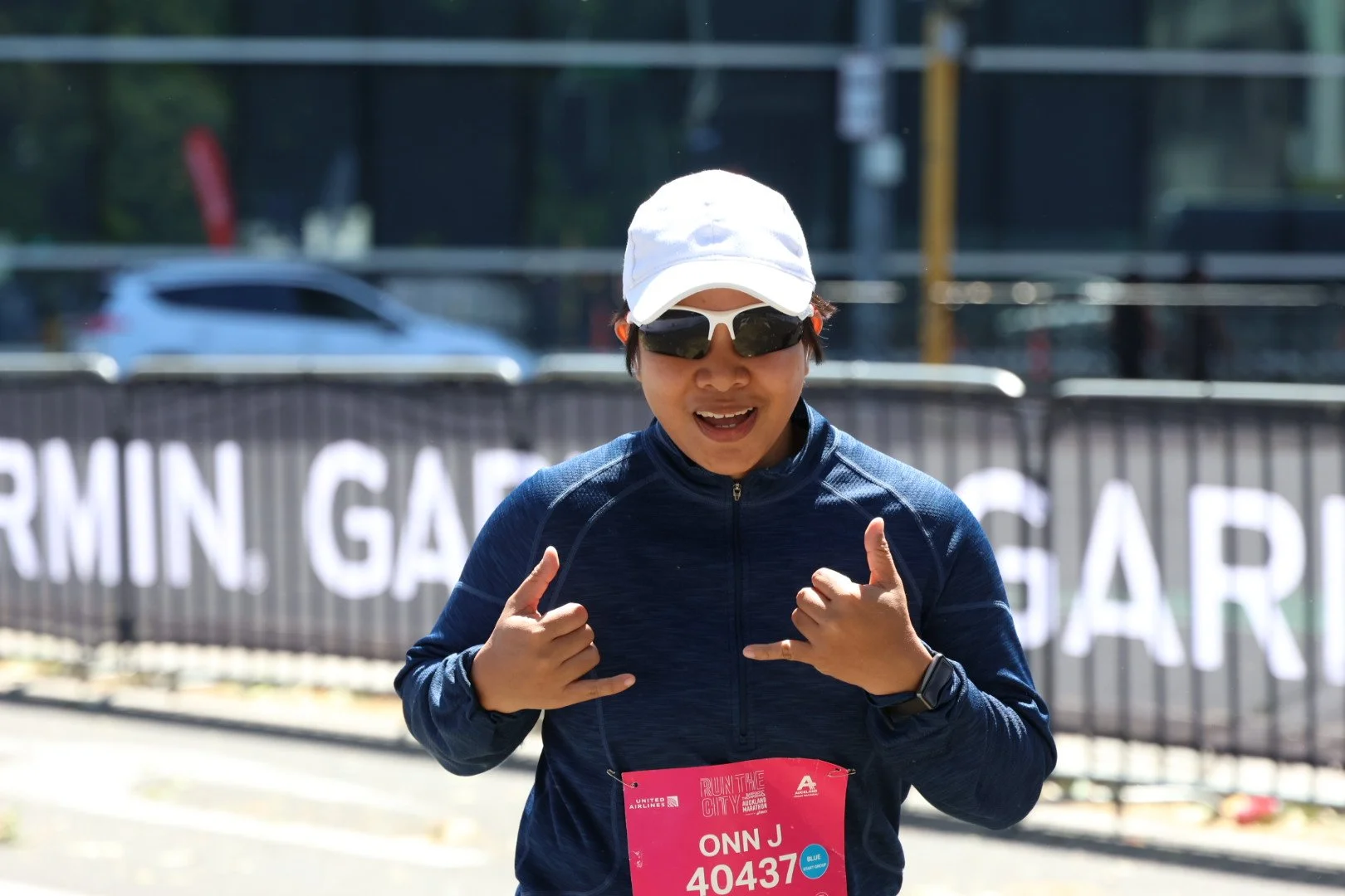 A woman in athletic gear and sunglasses smiling and making a gesture with her hands while participating in a running event.