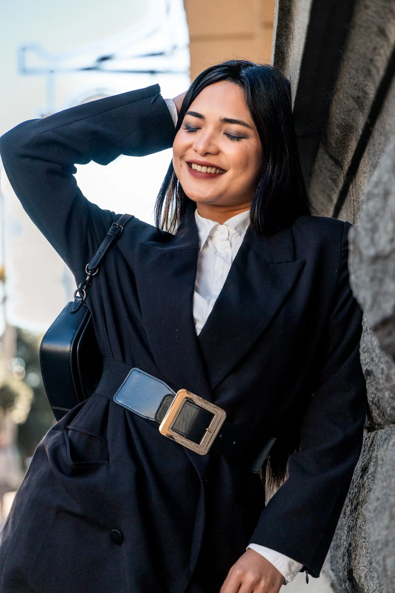 A woman with dark hair, smiling with eyes closed, leaning against a stone wall, wearing a black blazer, white shirt, and a large belt, holding a black handbag.