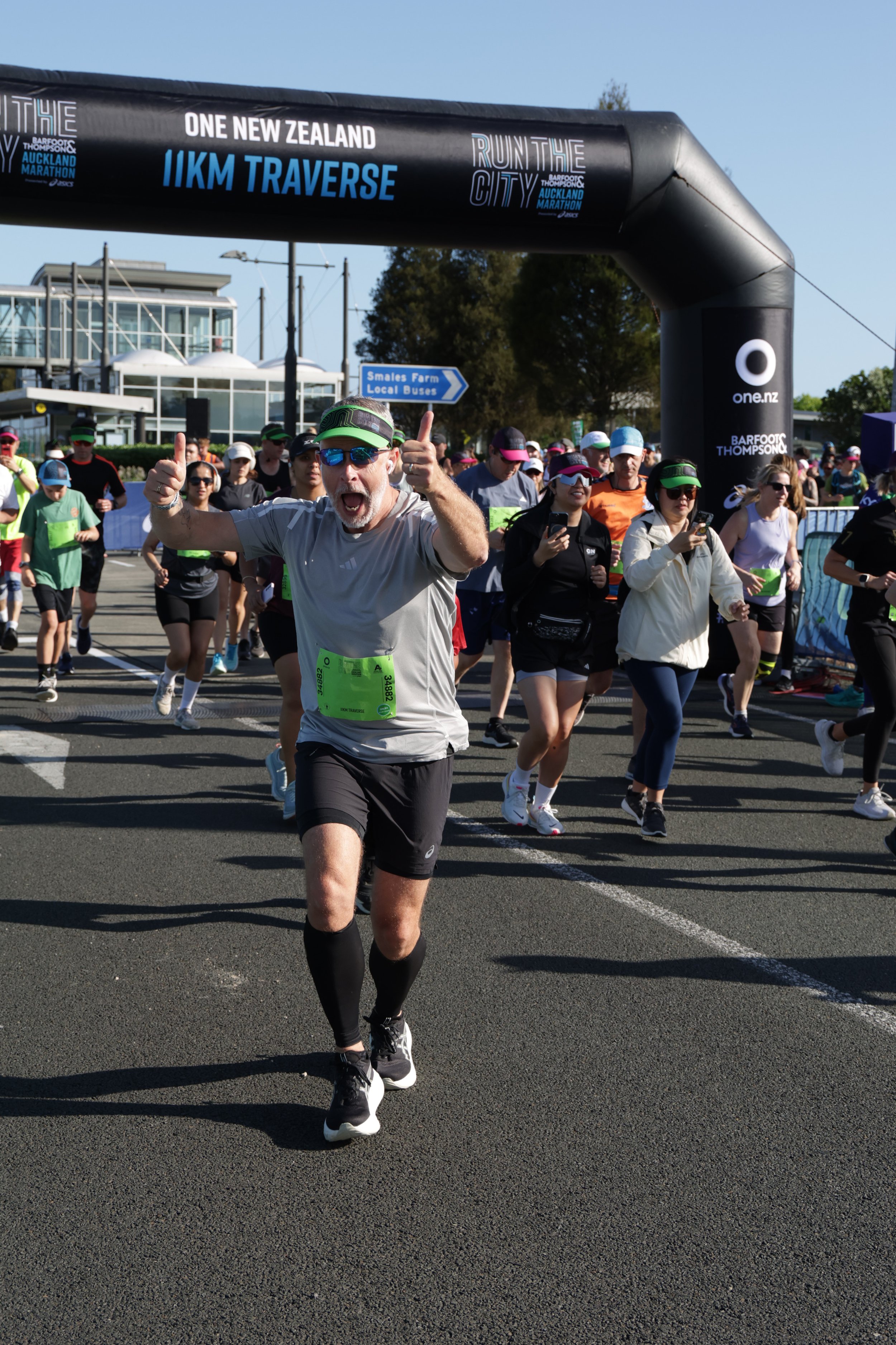 A man with a gray shirt and black shorts running in a marathon, giving a thumbs up, with many other runners behind him near a race arch. The race arch displays the text 'Run the City' and 'IIM Traverse' and is sponsored by one.nz.