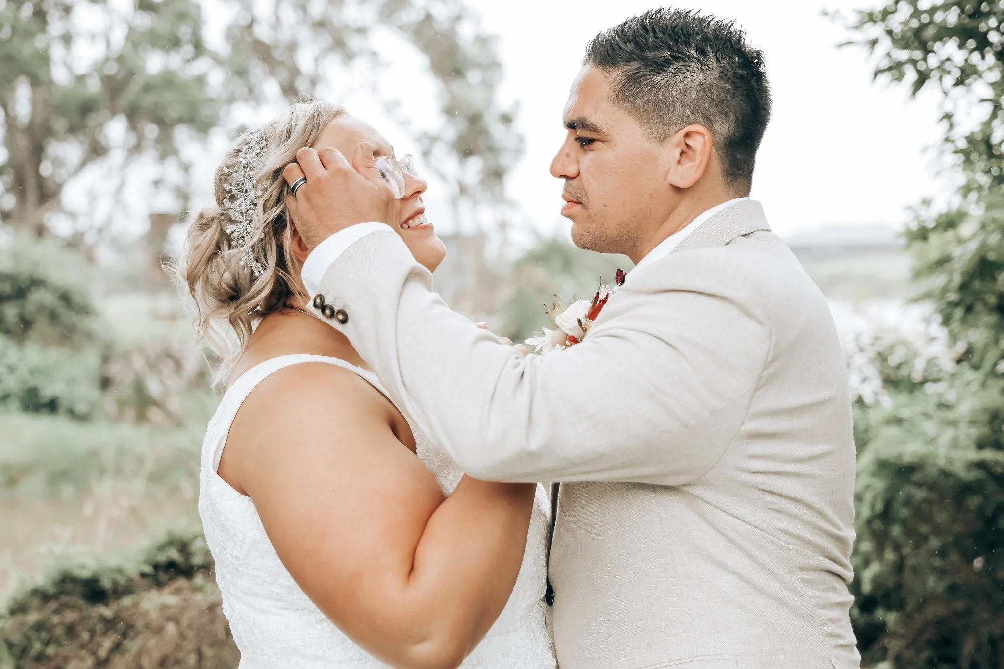 A bride and groom share a joyful moment outdoors, with the groom touching the bride's face and both smiling at each other.