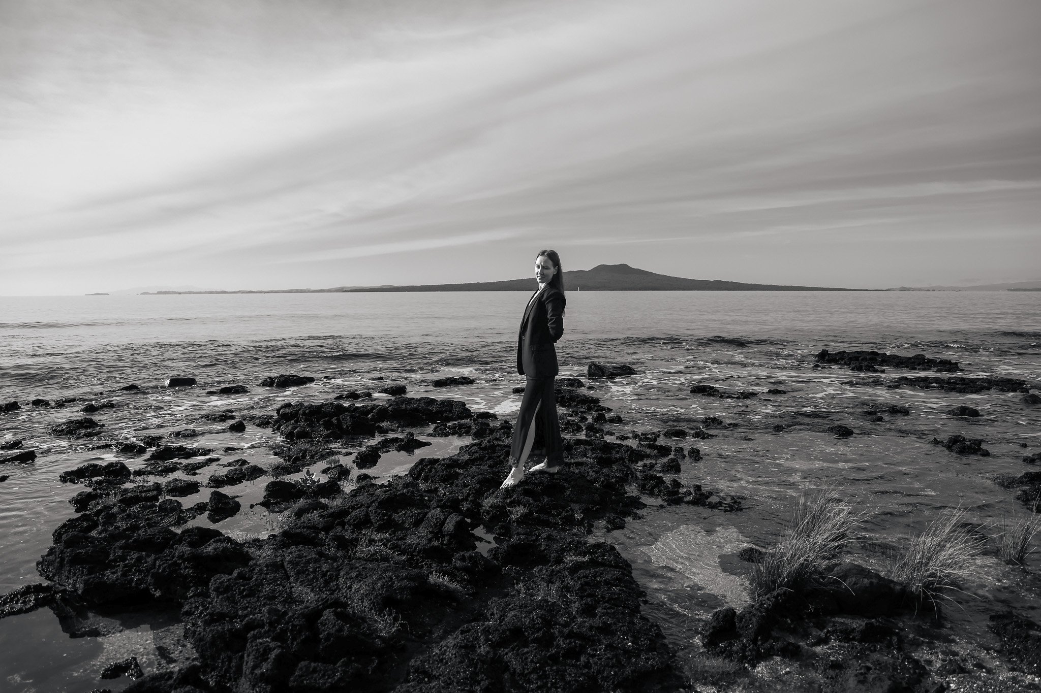 A woman in a coat standing on a rocky shoreline by the ocean with an island and a mountain in the background, captured in black and white.