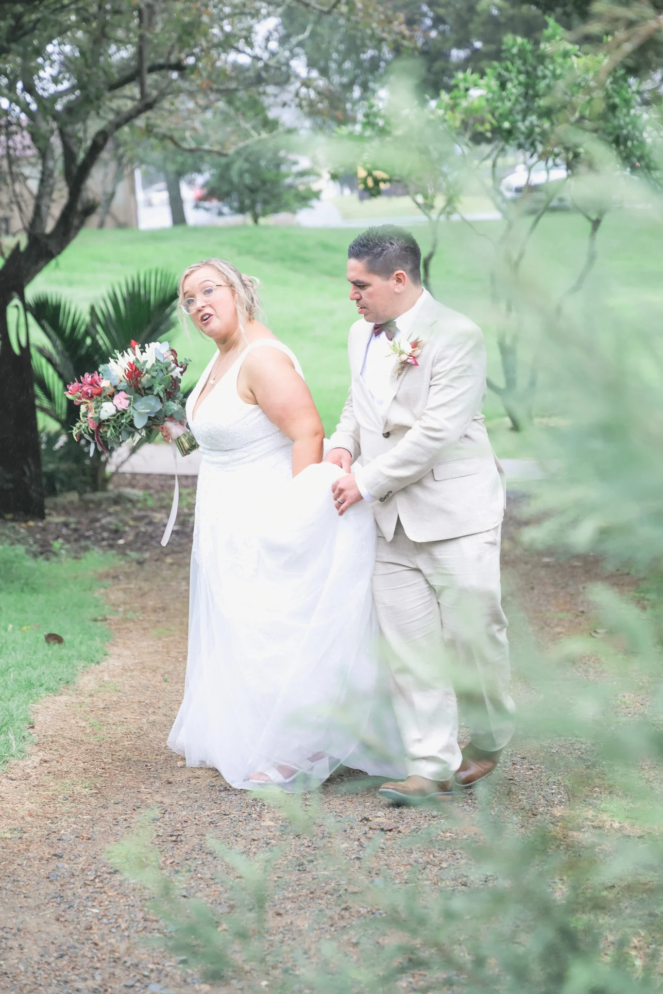 A bride and groom holding hands during their outdoor wedding, with the bride holding a bouquet of colorful flowers, surrounded by greenery.