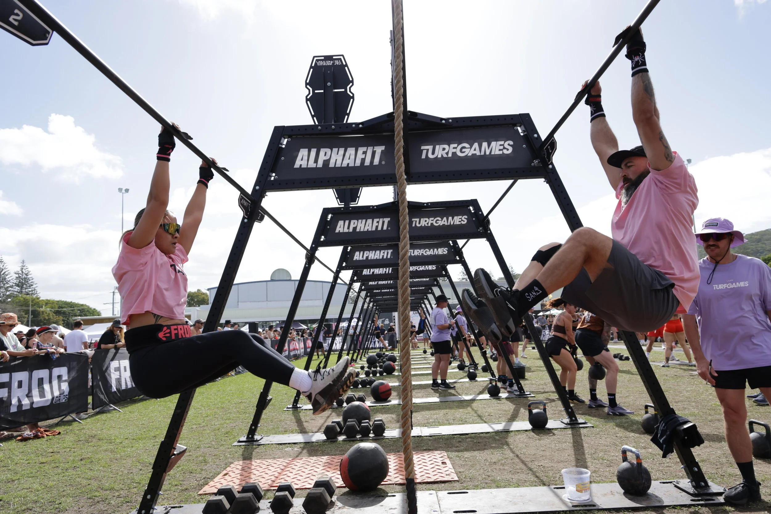 People participating in a strongman competition outdoors, lifting large black metal frames with weights, under a bright sky with clouds, surrounded by onlookers and event tents.