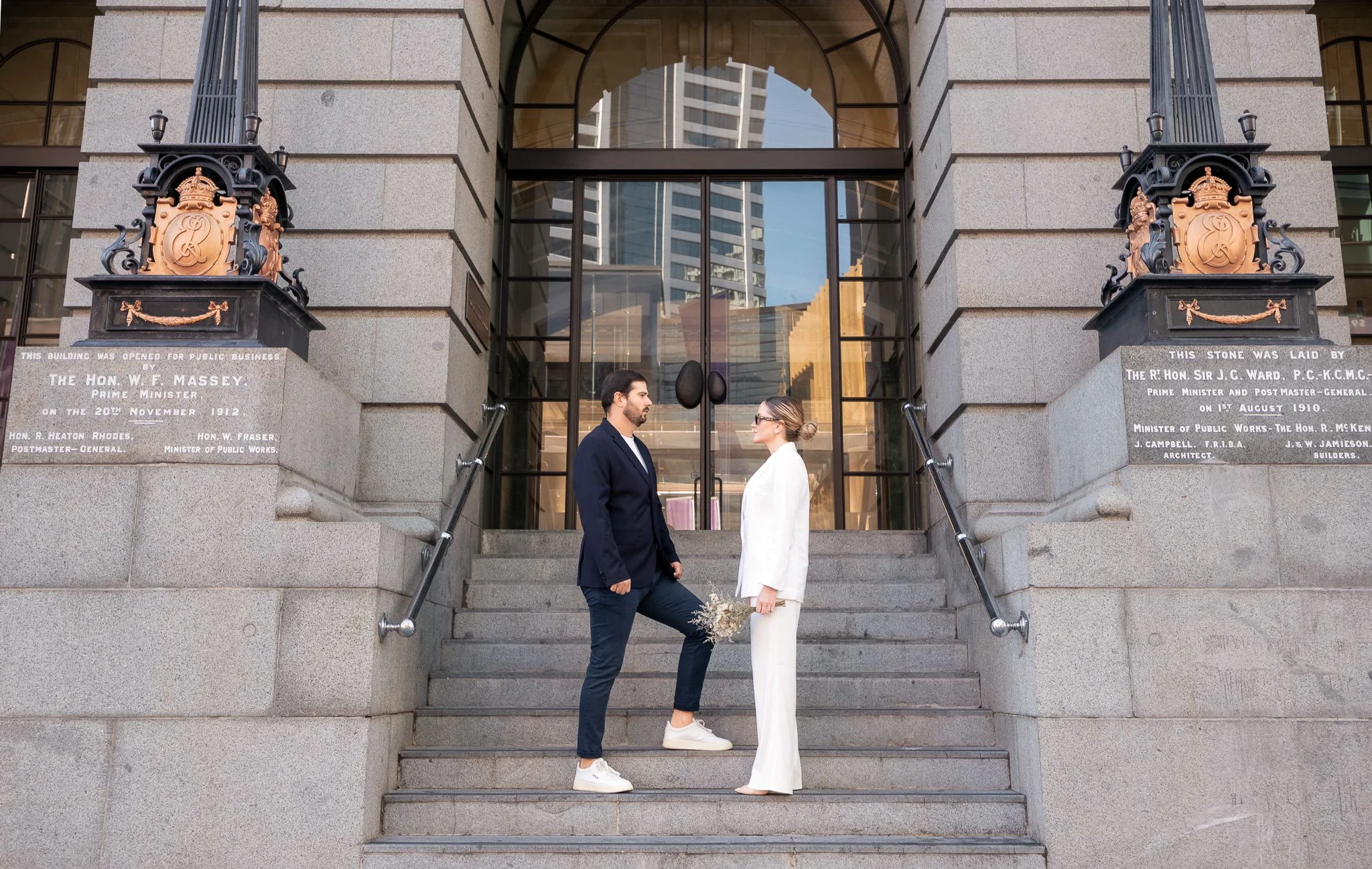 A man and woman standing on stone steps in front of a building with glass doors, facing each other, with the woman holding a bouquet of flowers and the man having one knee raised.