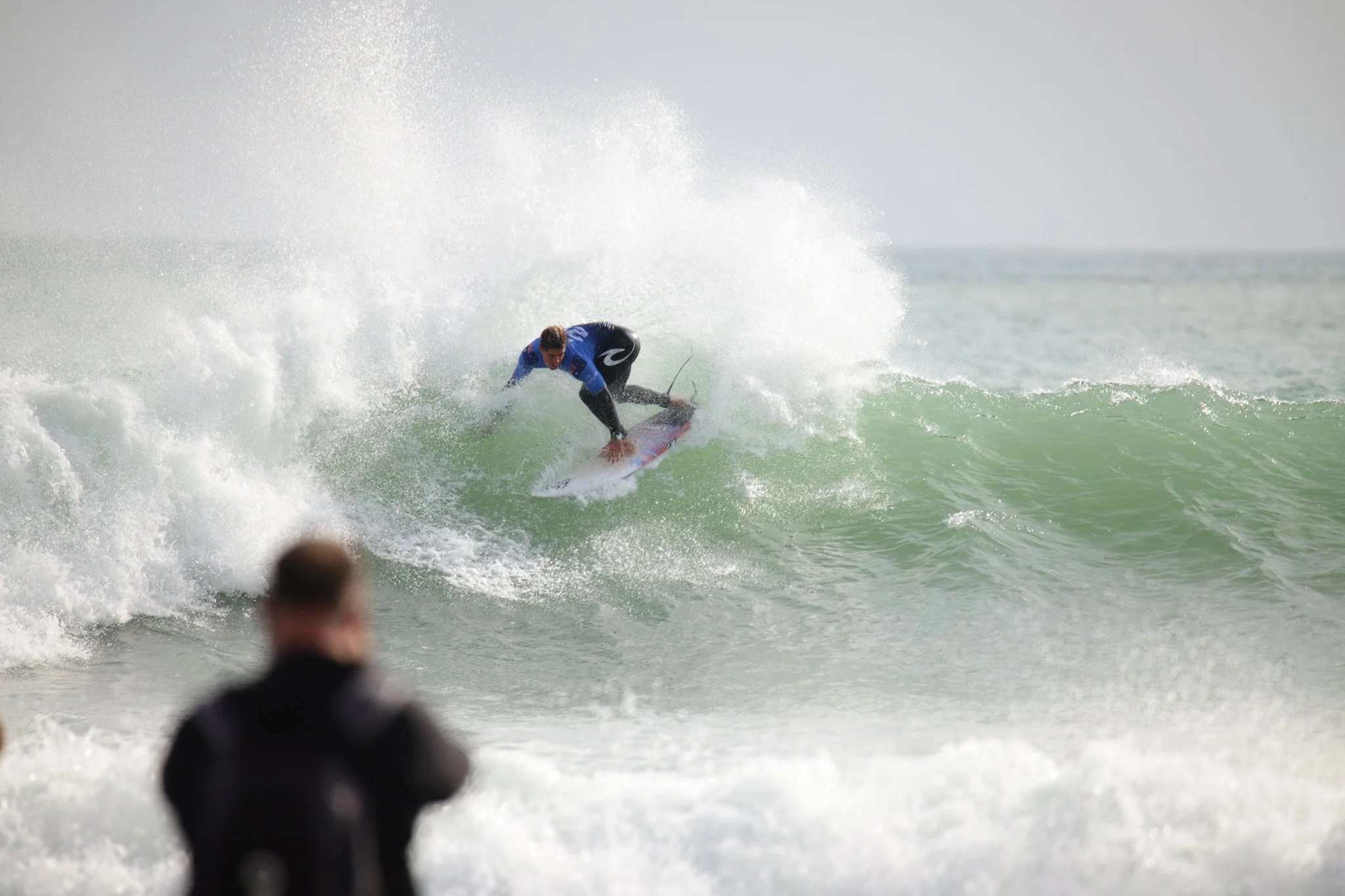 A surfer riding a wave at the beach, with spectators watching in the foreground.