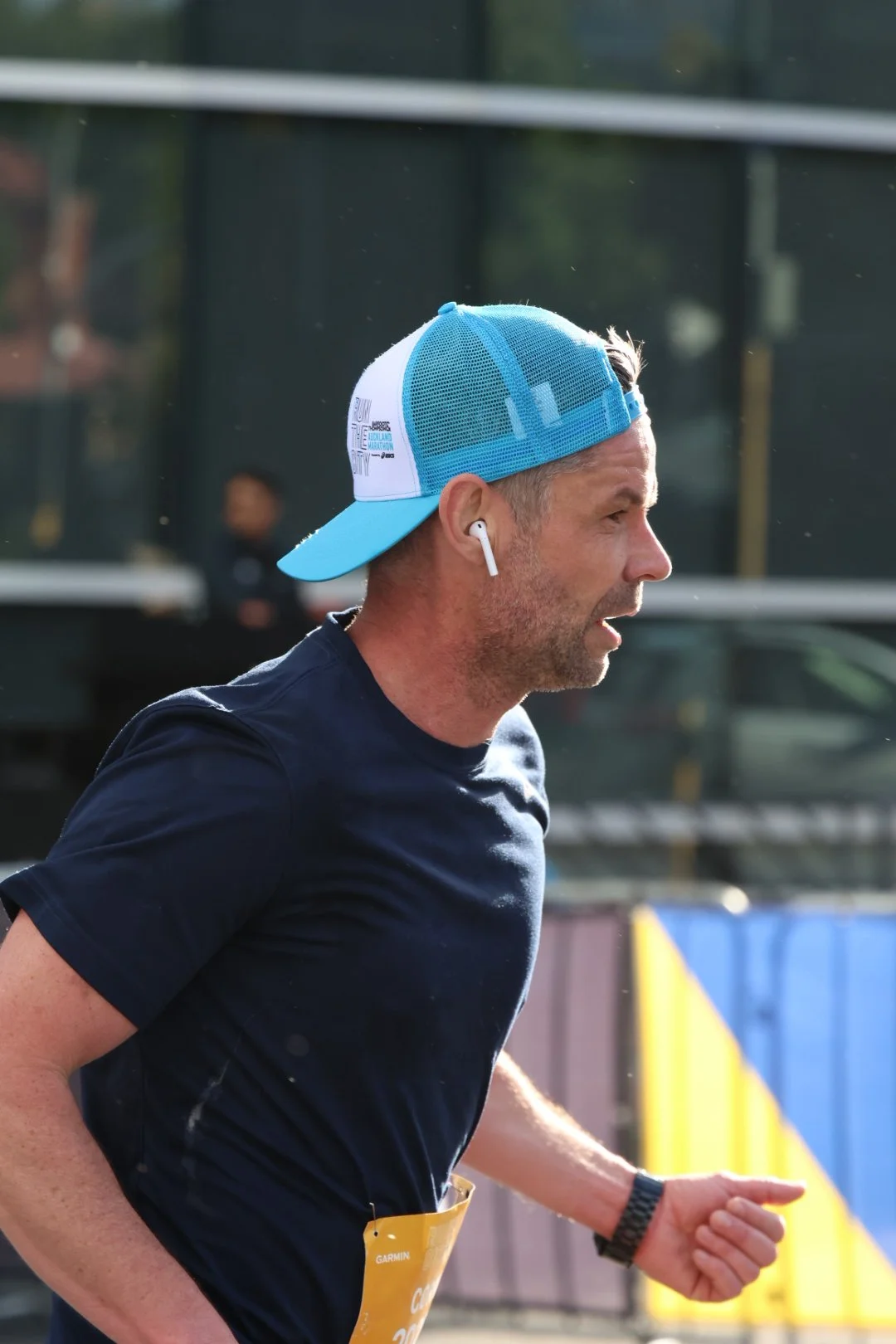 Man running during a race, wearing a blue and white trucker hat, black athletic clothing, and wireless earbuds.