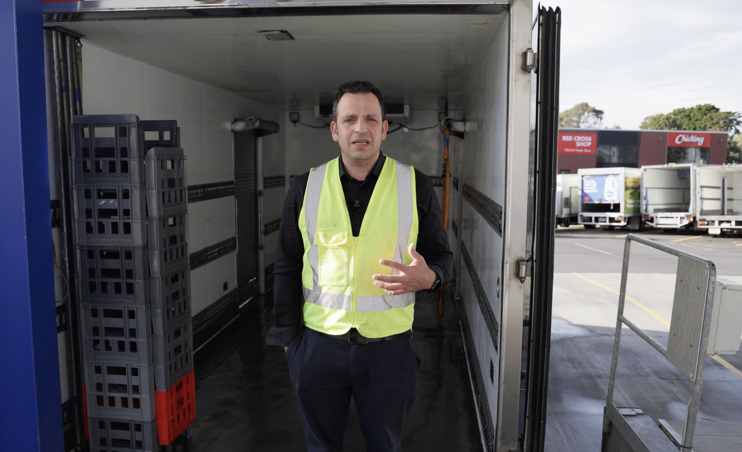 A man wearing a black shirt and a yellow safety vest standing inside an open truck trailer, with a parking lot and store in the background.