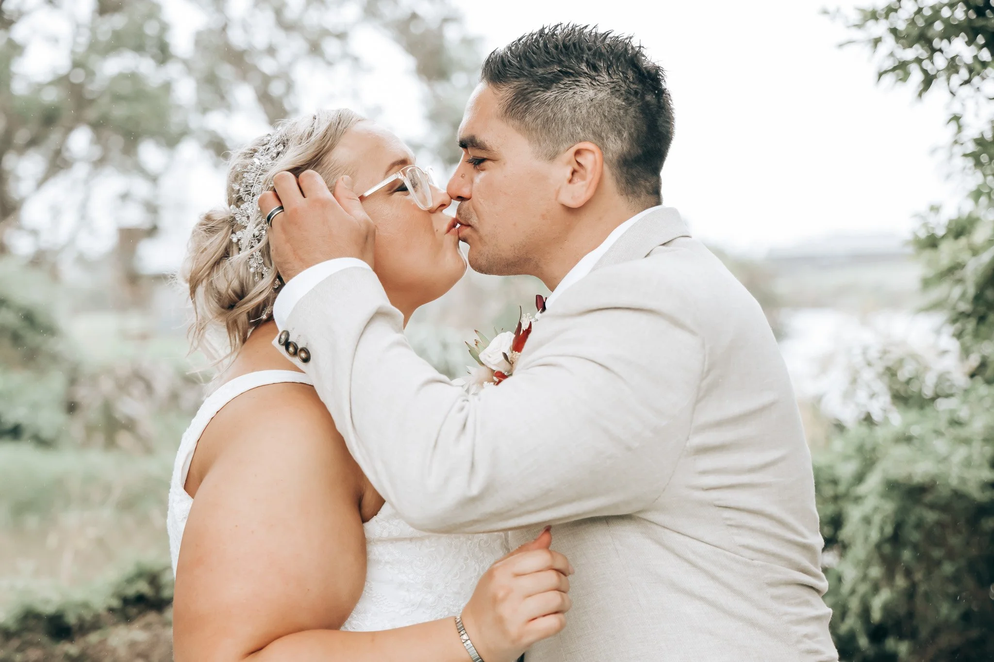 A bride and groom are kissing outdoors, holding each other's faces, with trees and greenery in the background.