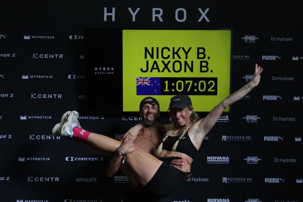 Two smiling athletes celebrating after a fitness competition, with one lifting the other. A large screen behind them displays their names, Nicky B. and Jaxon B., their time of 1:07:02, and an Australian flag. The background has various fitness and su