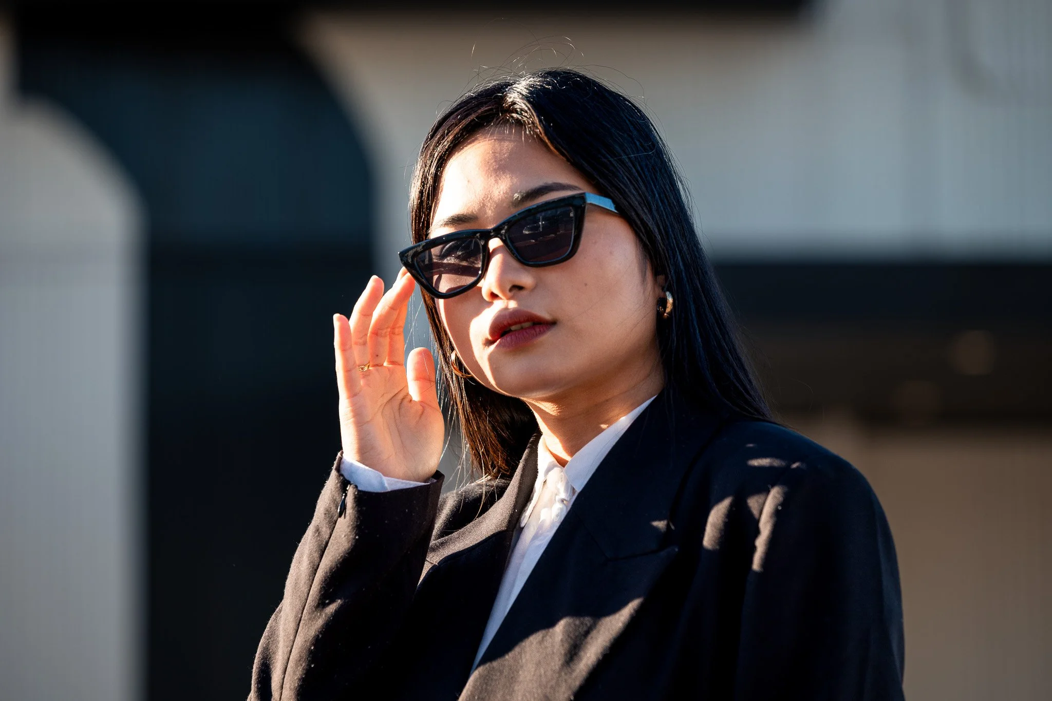 A woman with dark hair wearing sunglasses, a black blazer, and white shirt, outdoors during daytime.