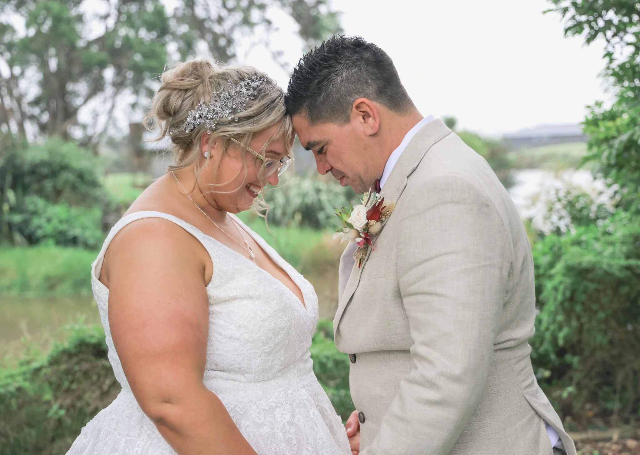 A bride and groom stand close together outdoors with heads touching and eyes closed, appearing to share a tender moment during their wedding. The bride has blonde hair styled with a decorative hairpiece and wears glasses and earrings. The groom has s