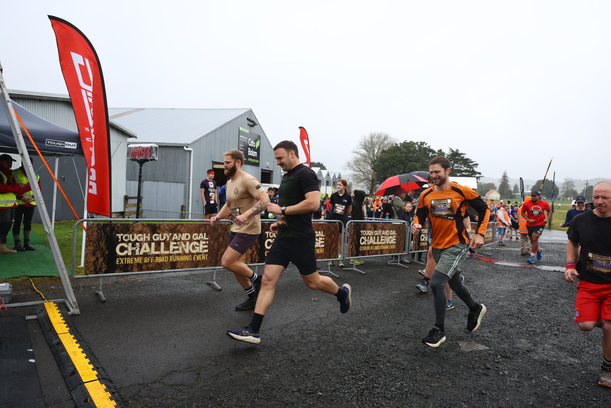 Participants nearing the finish line at the Tough Guy and Gal Challenge, an extreme off-road running event, on a rainy day with spectators and event staff around.