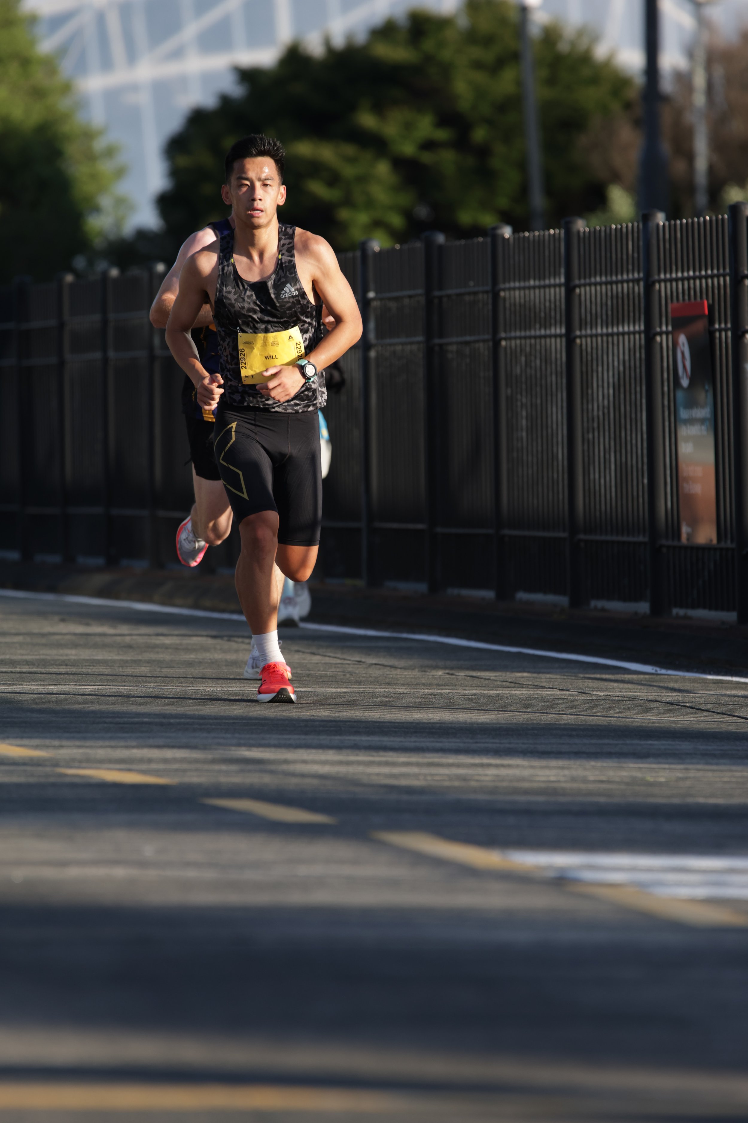 A male runner in a black and gray athletic uniform running on a paved road with a black fence and green trees in the background.