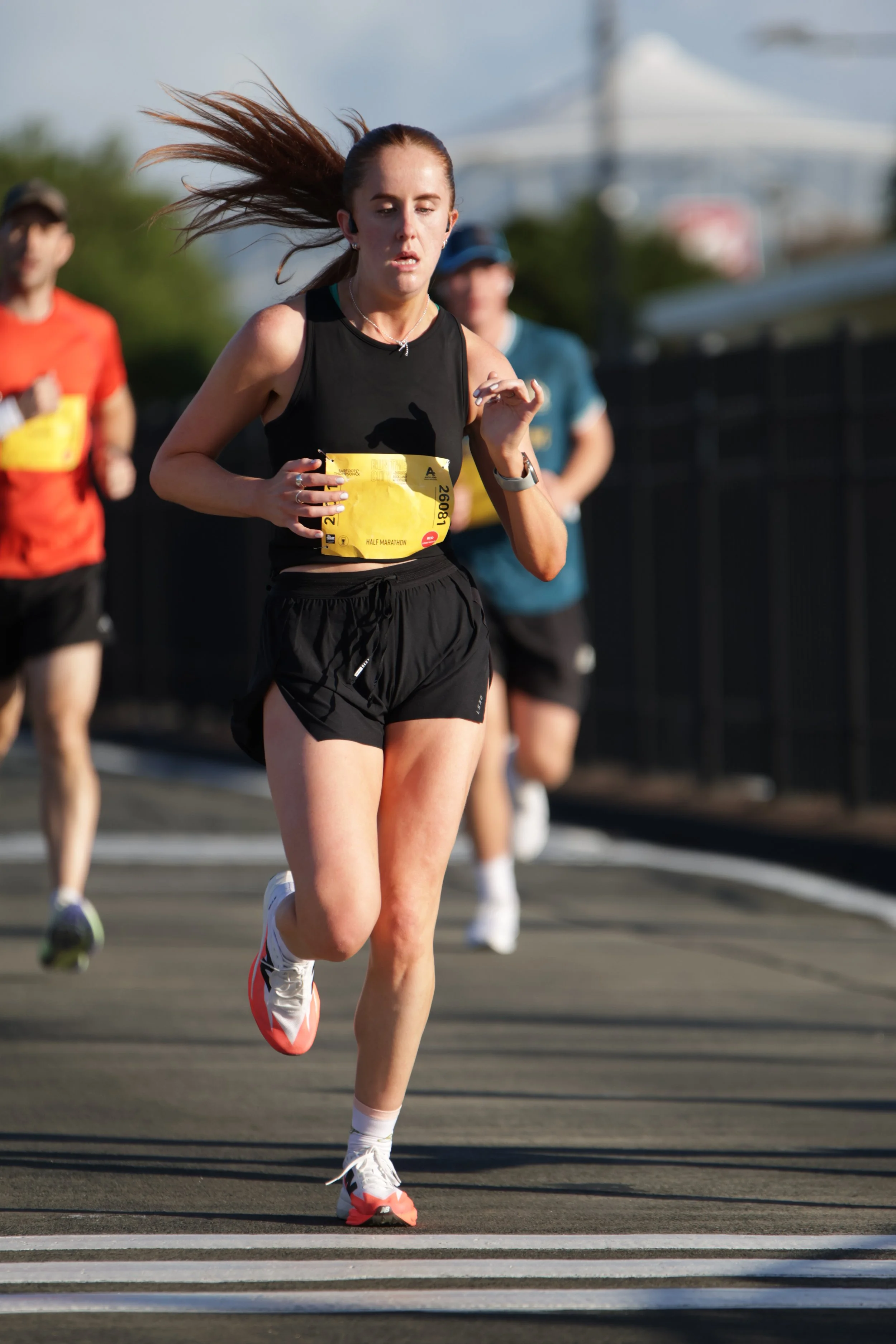 A woman running in a race, wearing a black athletic top, black shorts, white and red running shoes, and a yellow race bib number 26081 on her waist, with other runners in the background.