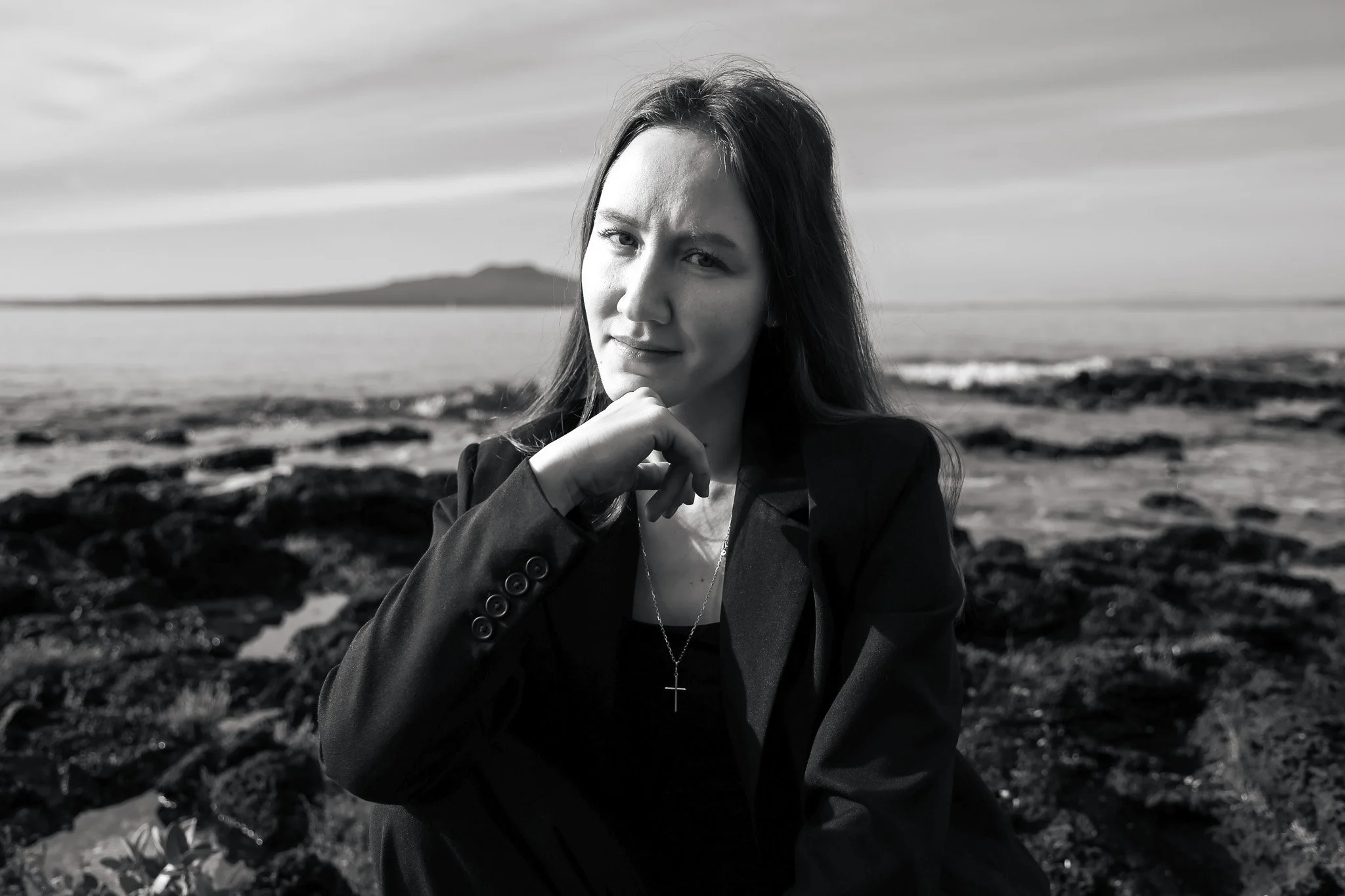 A woman with long hair, wearing a blazer and a cross necklace, posing outdoors near the beach or rocky shoreline with mountains in the background.